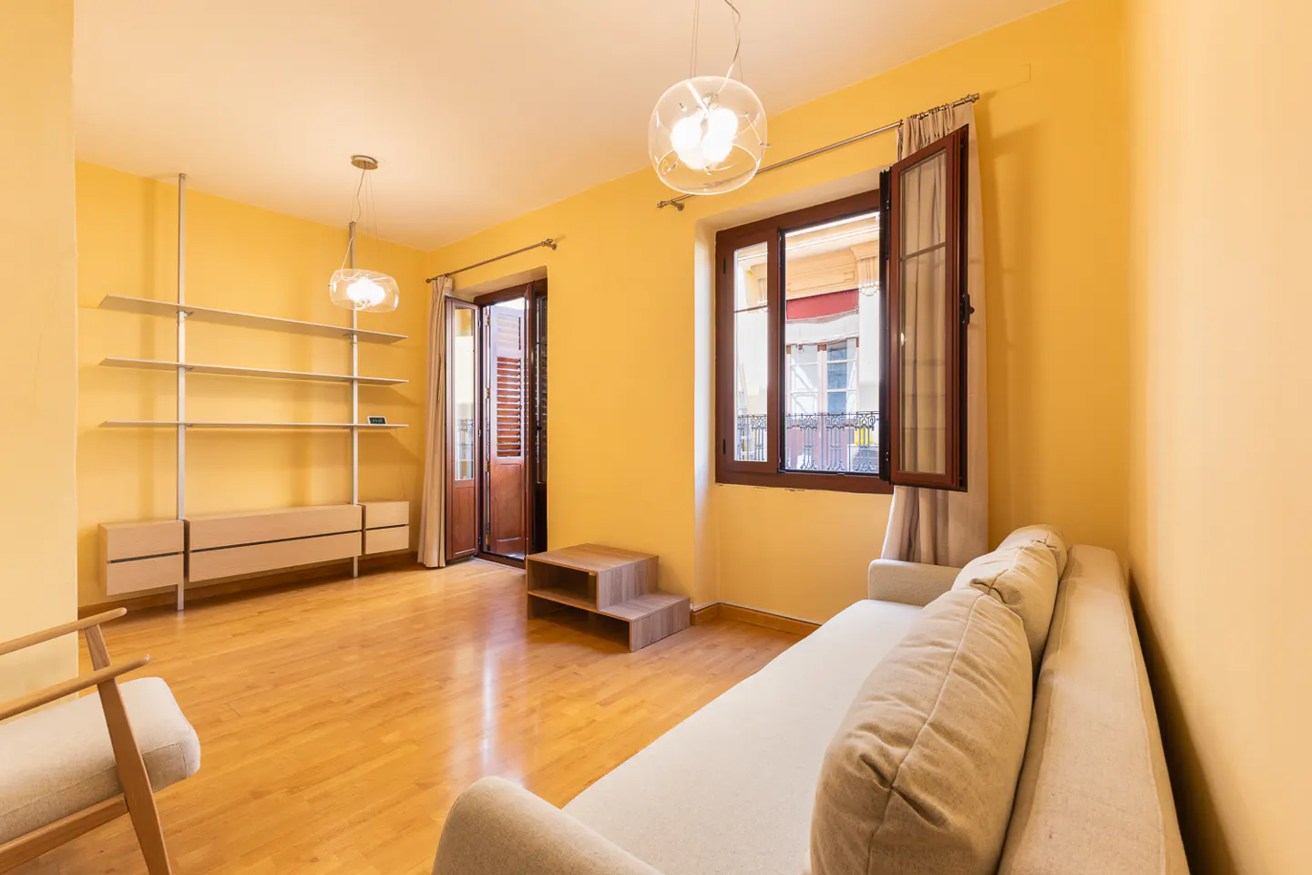 Bright living room with yellow walls, light wood floors, a beige sofa, open shelving, and a window with wooden shutters.