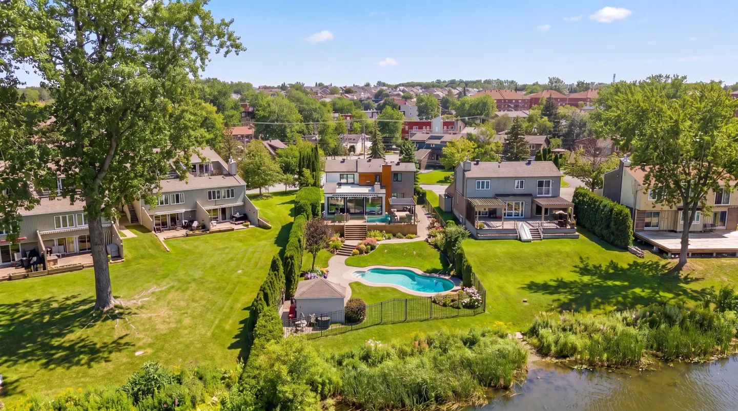 Aerial view of houses with green lawns, a kidney-shaped pool, and a lake in the foreground on a sunny day.