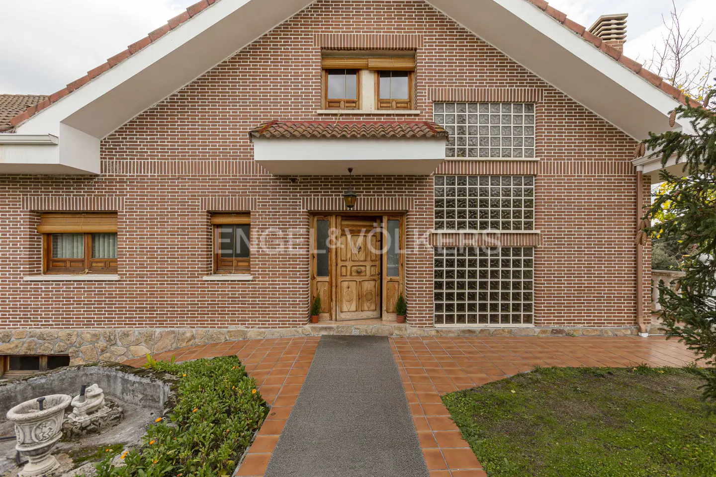 Exterior of a two-story brick house with a wooden door, glass block window, and a gray walkway.