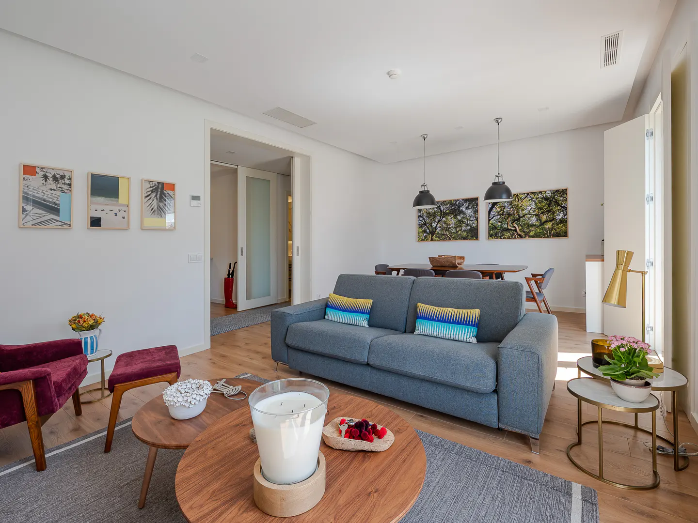 Bright living room with a gray sofa, wood floors, and white walls. Art hangs on the walls, and a dining table is visible in the background.