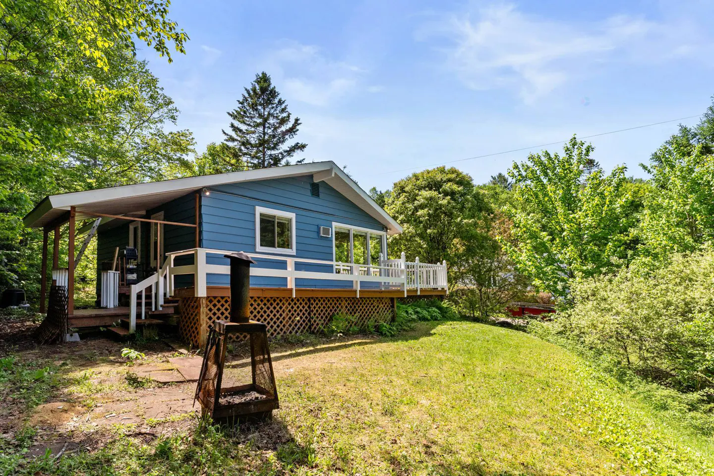 Blue cottage with a white deck and lattice skirting, set in a lush, sunny backyard with a metal fire pit in the foreground.