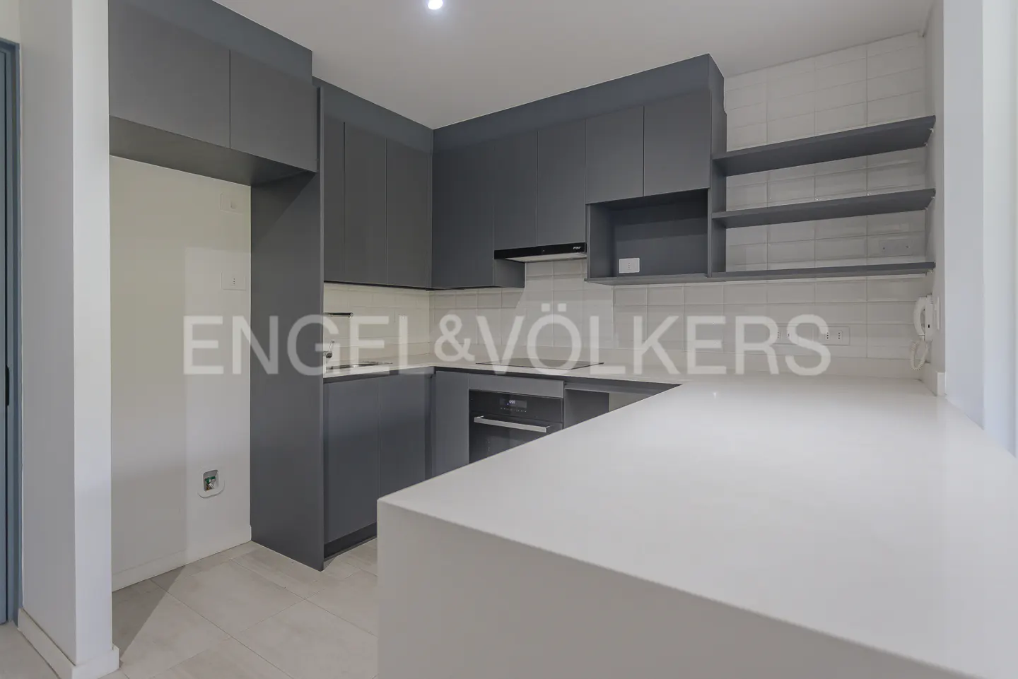 A modern kitchen with gray cabinets, white countertops, and white subway tile backsplash. Open shelving and stainless steel appliances are visible.