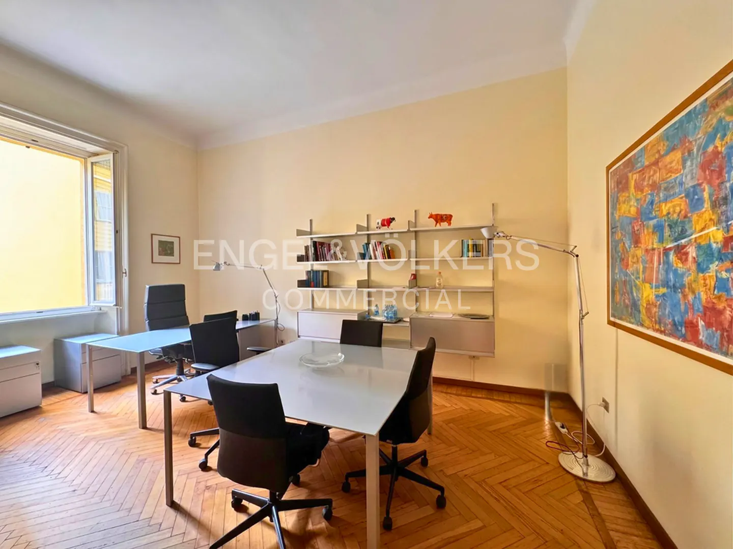 Office space with two white desks, black chairs, and a bookshelf. A colorful painting hangs on the wall. The floor is wooden parquet.