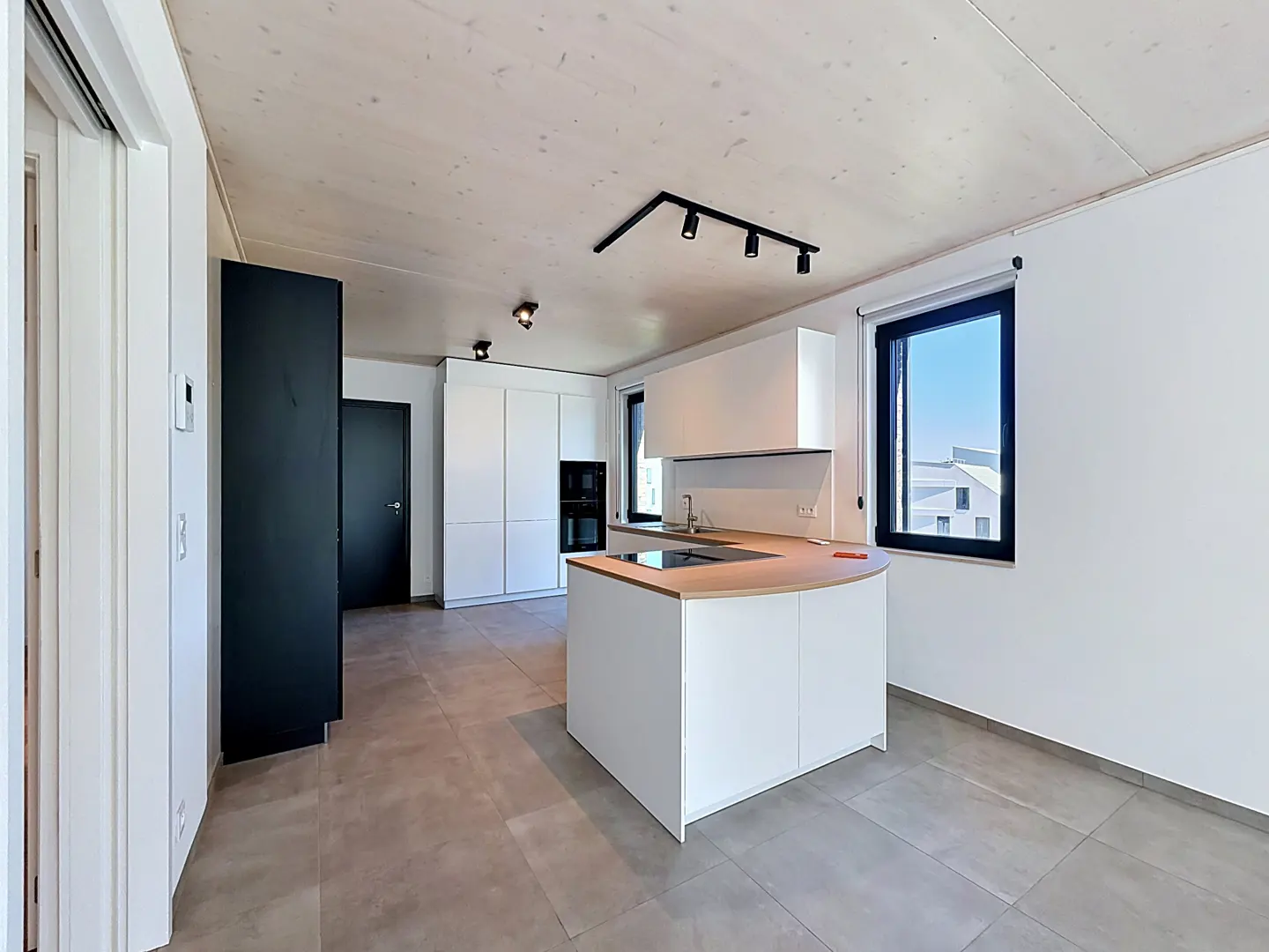 A modern kitchen with white cabinets, a curved island, and black accents. The room has gray tile flooring and a light wood ceiling.