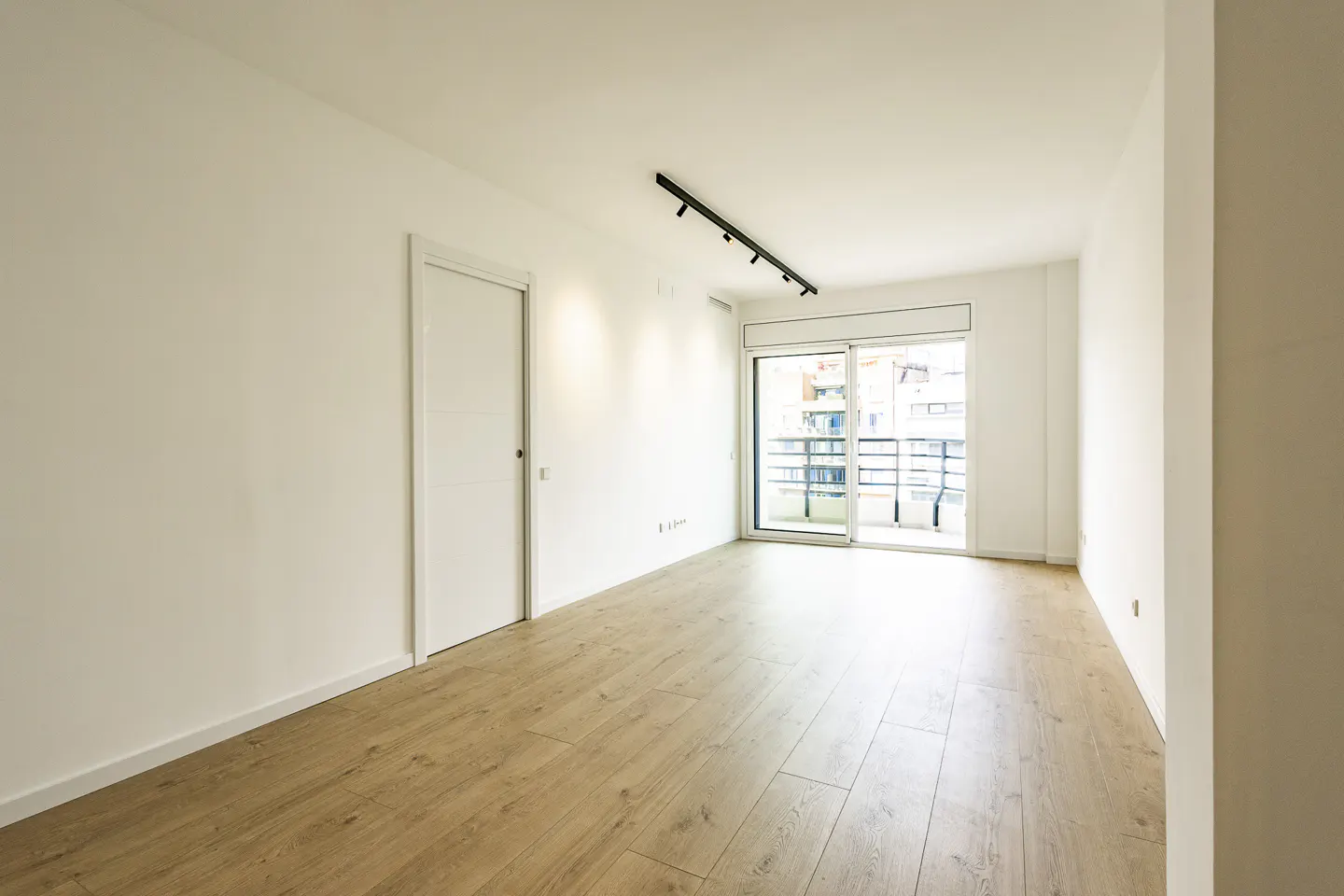 Bright, empty room with light wood floors, white walls, and a sliding glass door to a balcony. A black track light is on the ceiling.