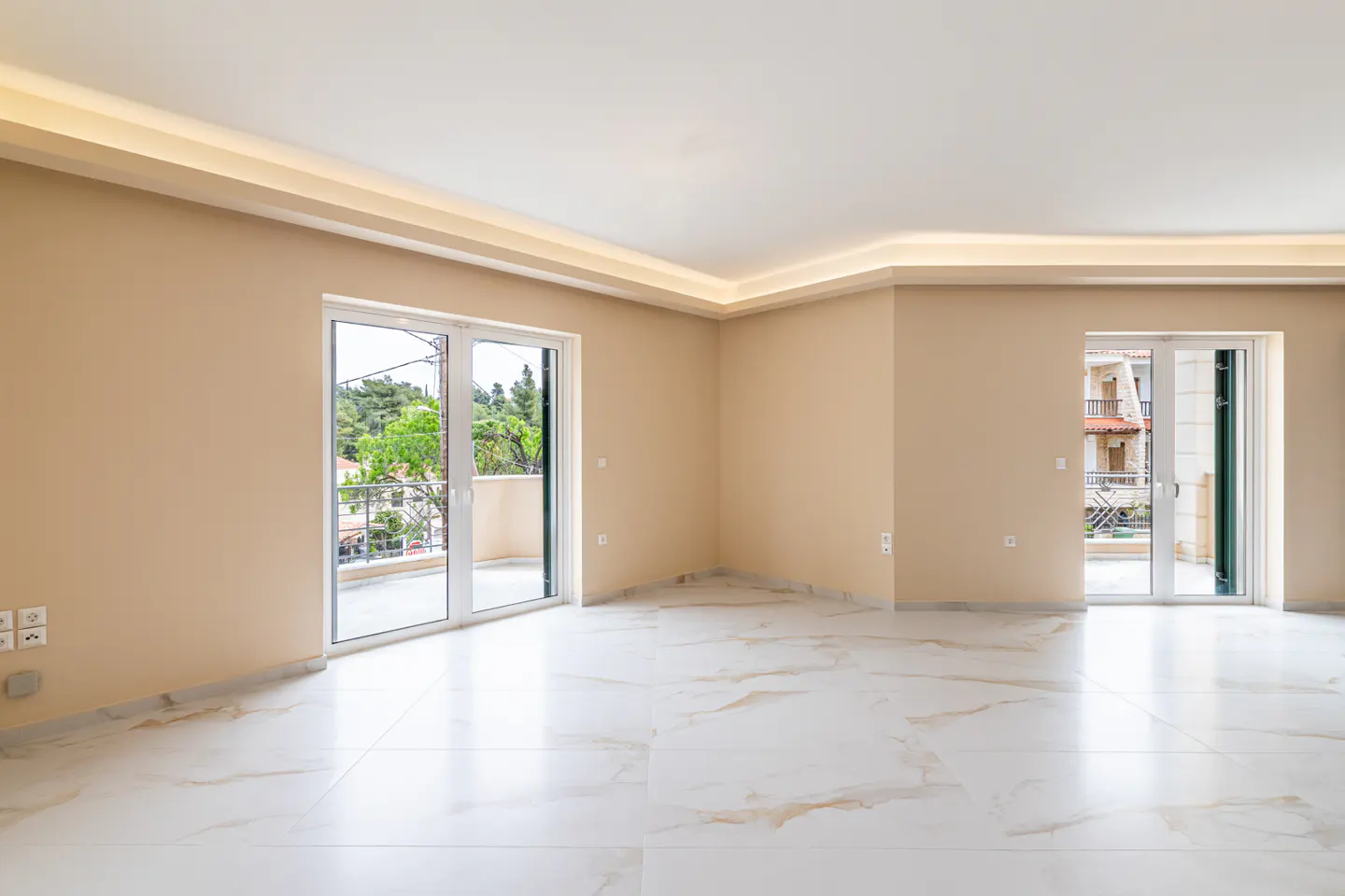Empty room with beige walls and white marble floor. Two sets of glass doors lead to balconies with views of trees and buildings.