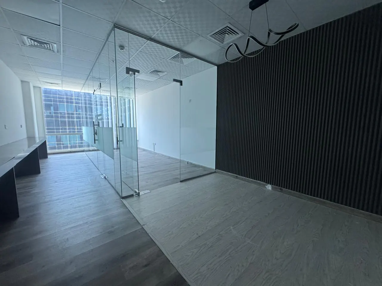 Empty office space with glass-walled rooms, gray wood floors, and a black accent wall with a modern light fixture.