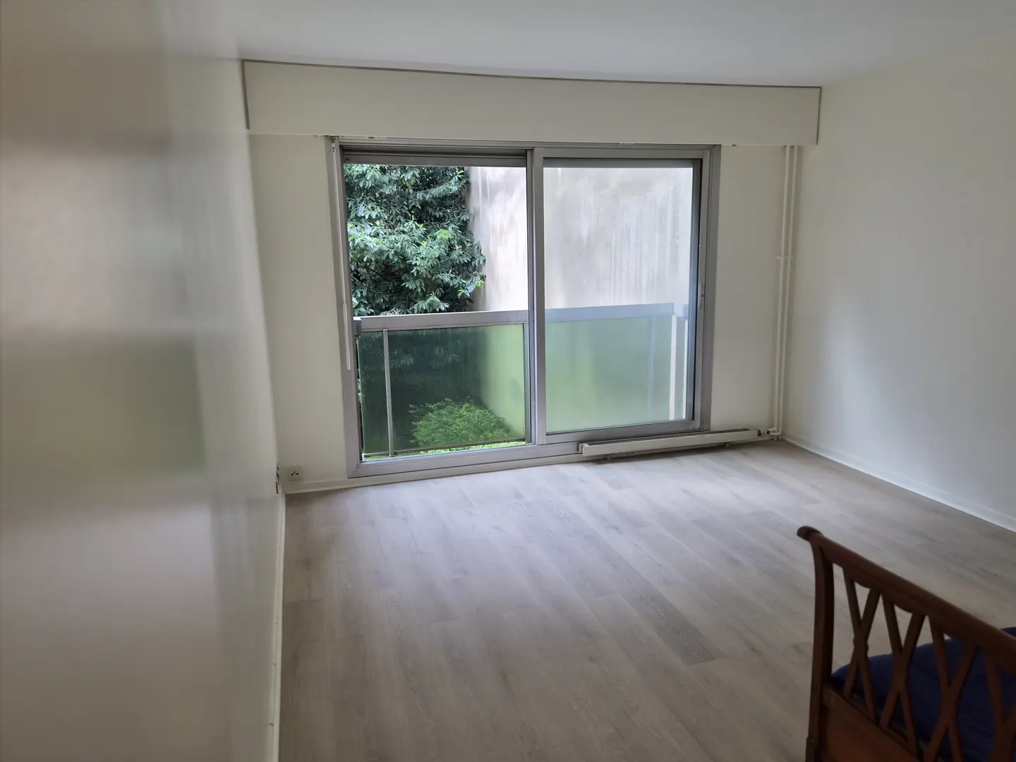 Empty room with light wood floors, white walls, and a large sliding glass door looking out onto green trees.