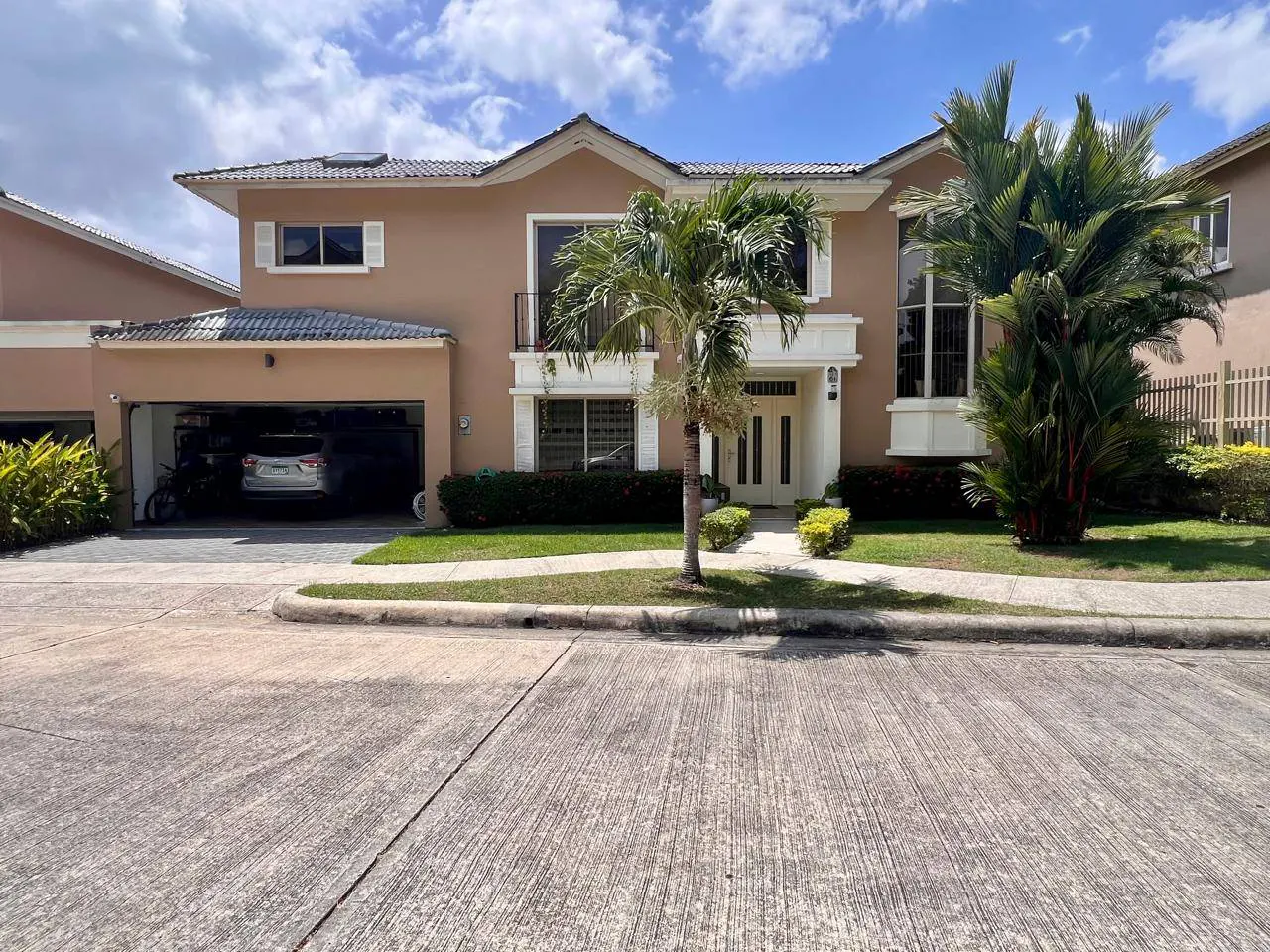Tan two-story house with white trim, a dark gray roof, and an open garage with a car inside. Palm trees and green grass in the front yard.