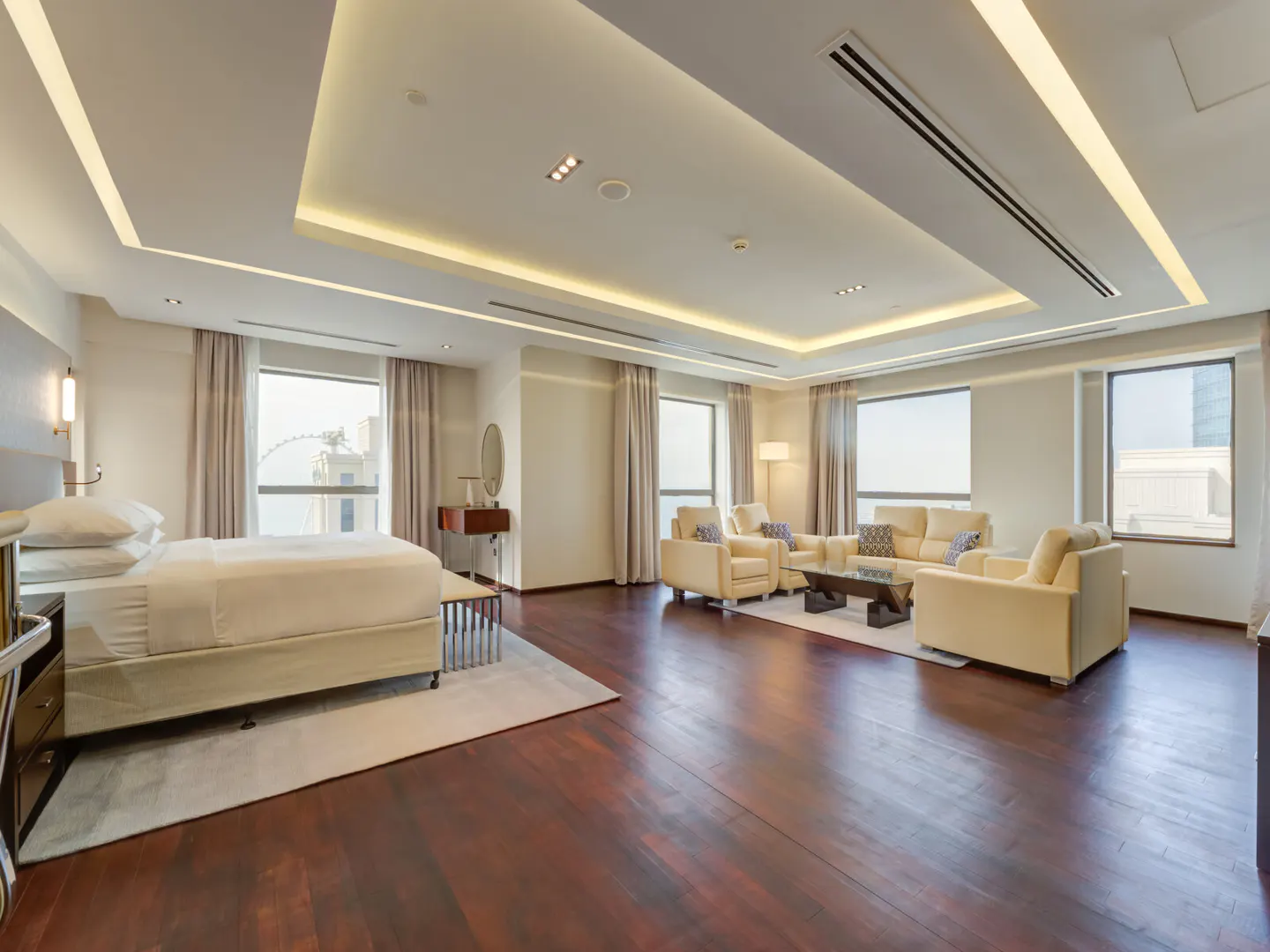 Bright, spacious suite with a king bed, sitting area with cream sofas, and dark wood floors. Large windows offer city views.