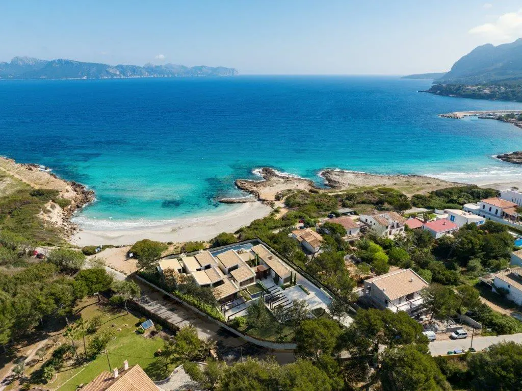 Aerial view of a modern, beige house with a pool near a turquoise ocean and sandy beach. Mountains are visible in the distance.