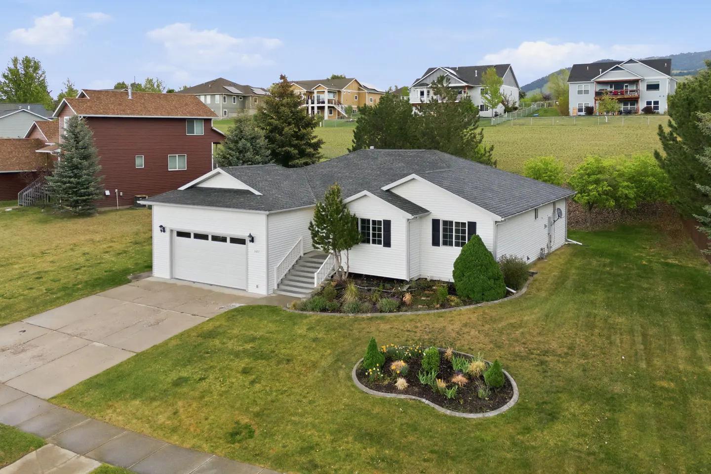 Aerial view of a white, single-story house with a gray roof, green lawn, and flower garden. Other houses are visible in the background.
