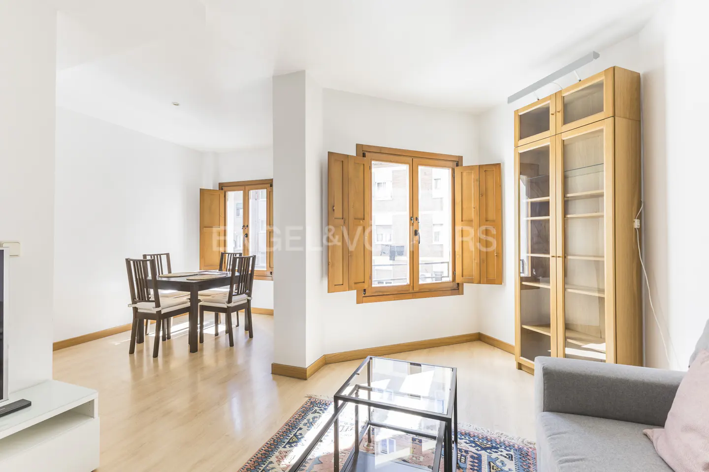 Bright living room with wood floors, white walls, and open windows with wood shutters. A dining table and chairs are visible in the background.