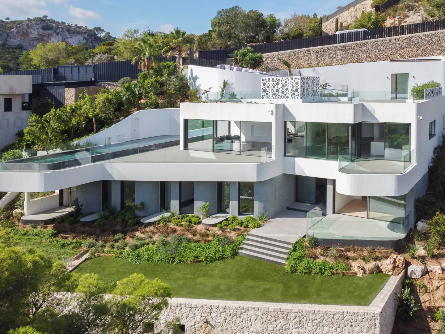 Modern white house with a pool on the roof, surrounded by green trees and a stone wall.