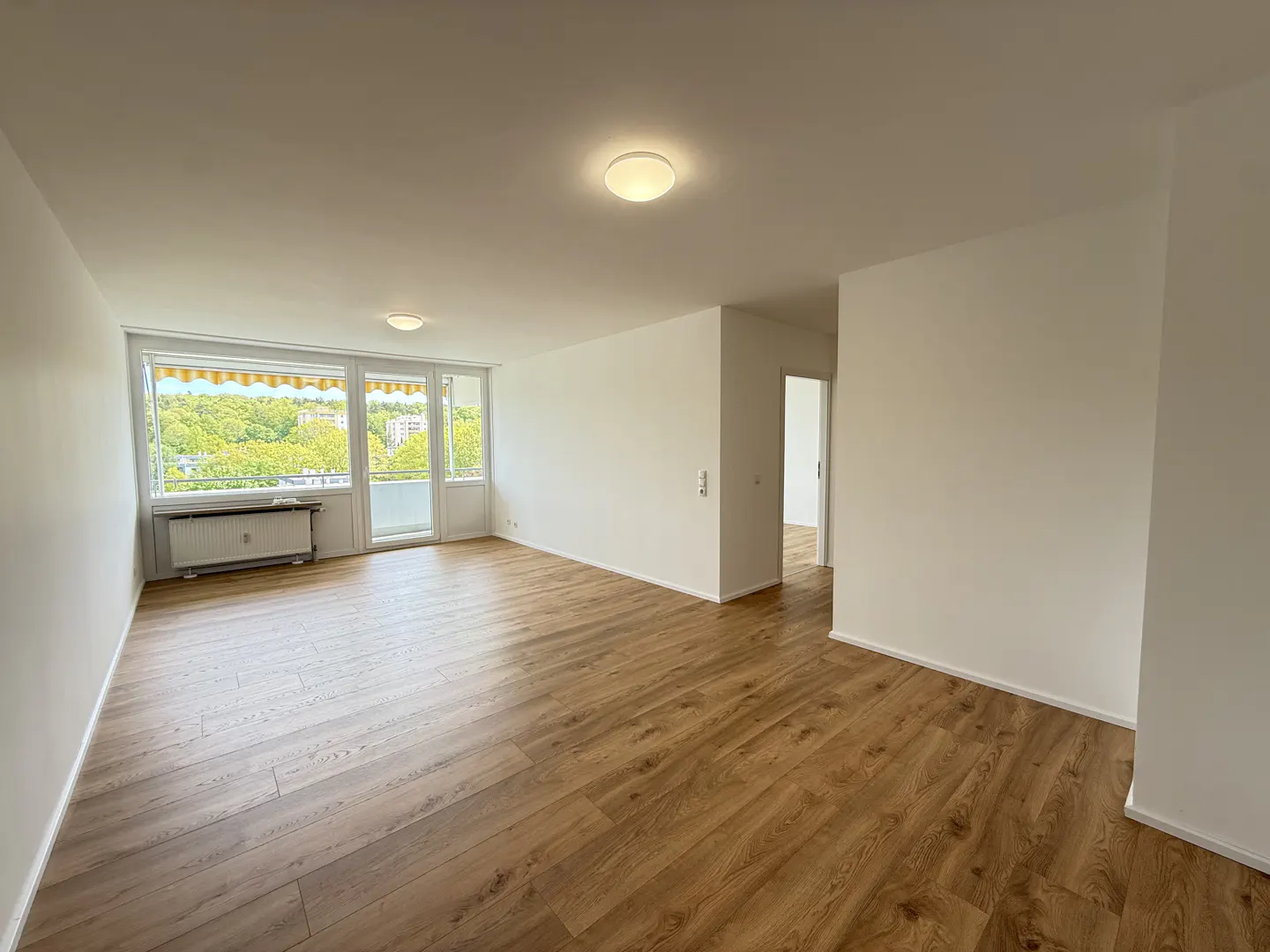 Spacious, empty living room with light wood-look flooring, white walls, and a large window overlooking green trees.