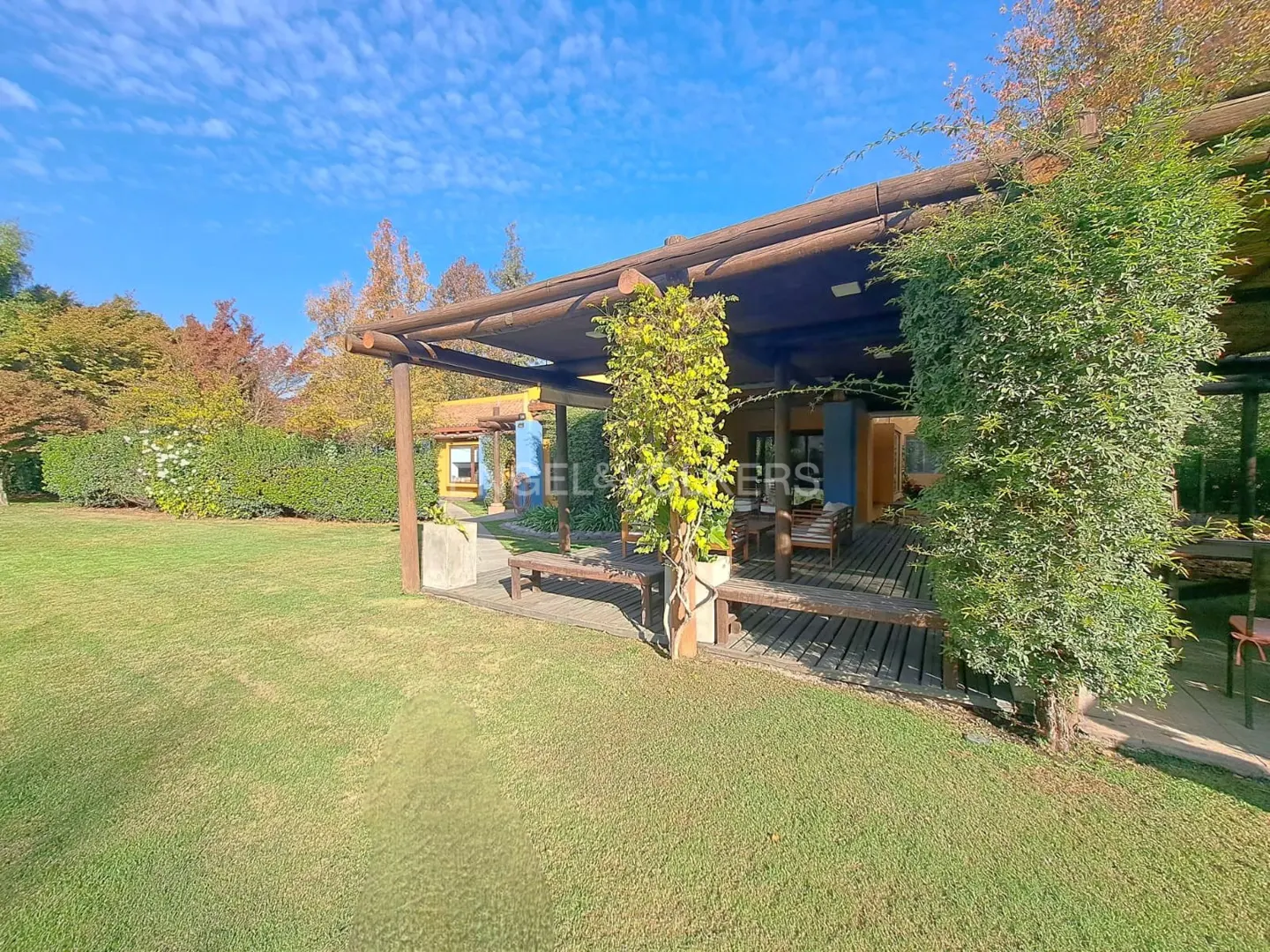 Lawn view of a house with a wooden patio and pergola covered in vines, under a blue sky.