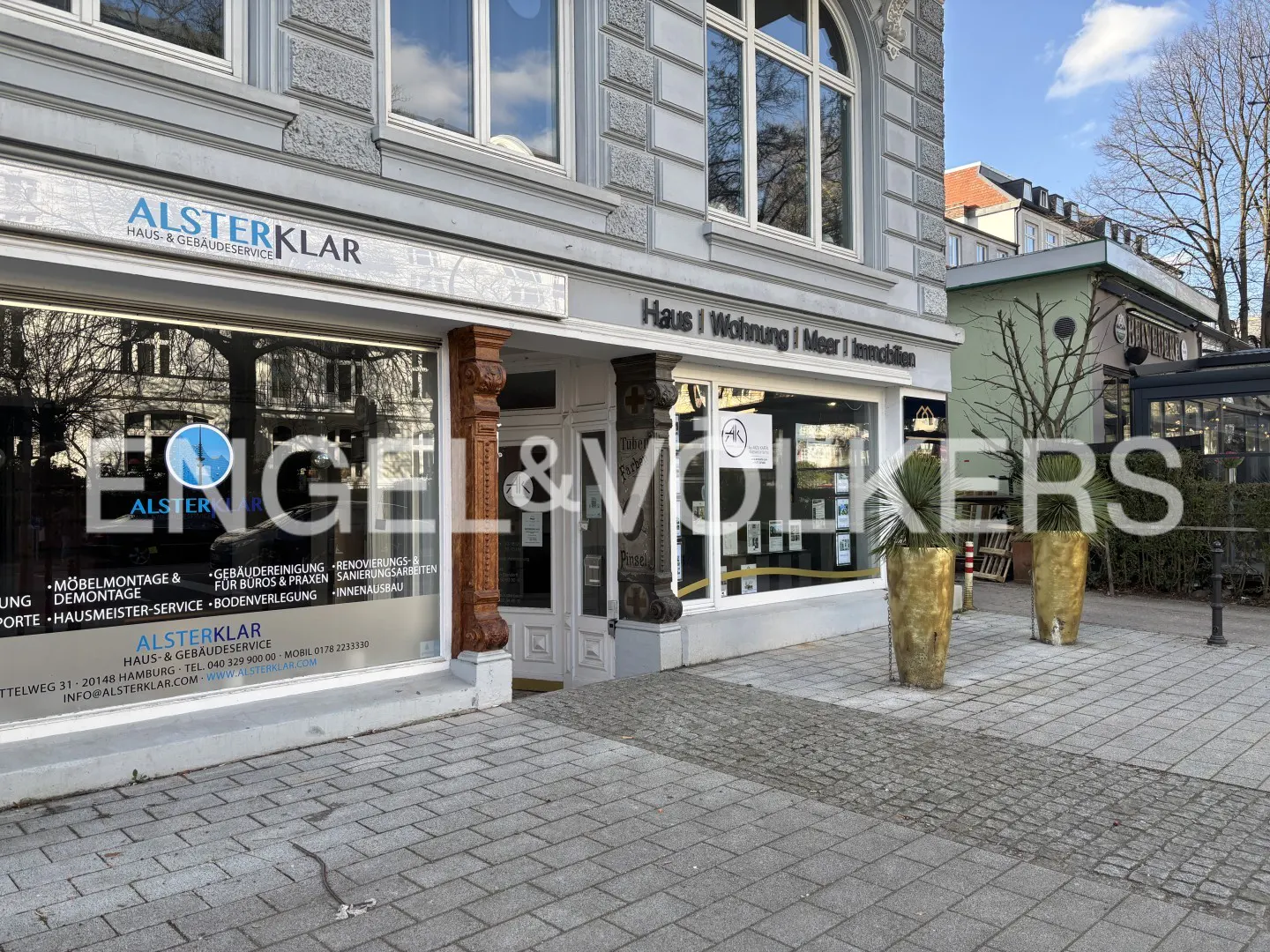 Exterior view of a grey commercial building with large windows, gold planters, and signage for Alsterklar and real estate.