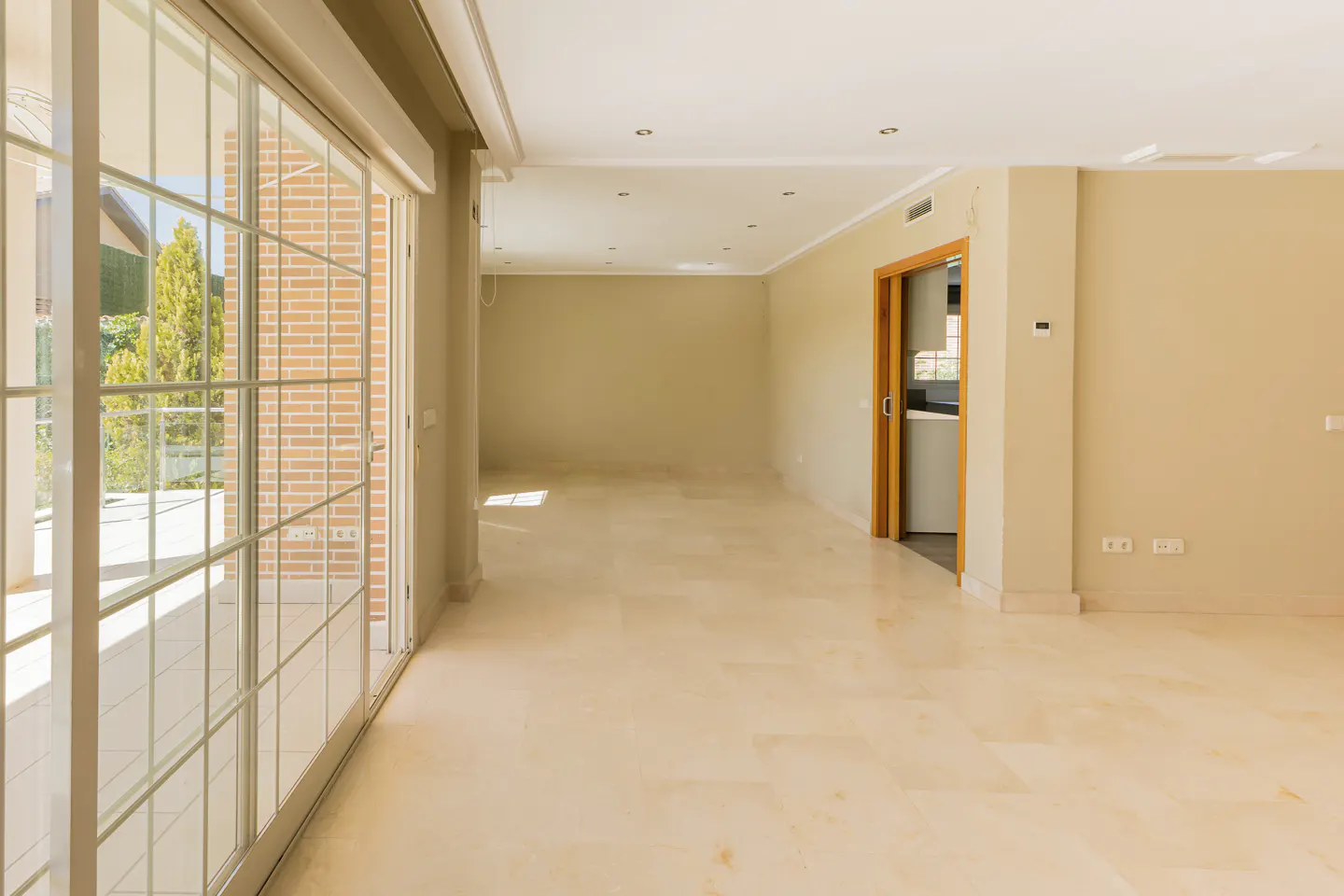 Bright, empty room with beige walls and marble floors. Large sliding glass doors lead to a patio. A doorway reveals a glimpse of a kitchen.