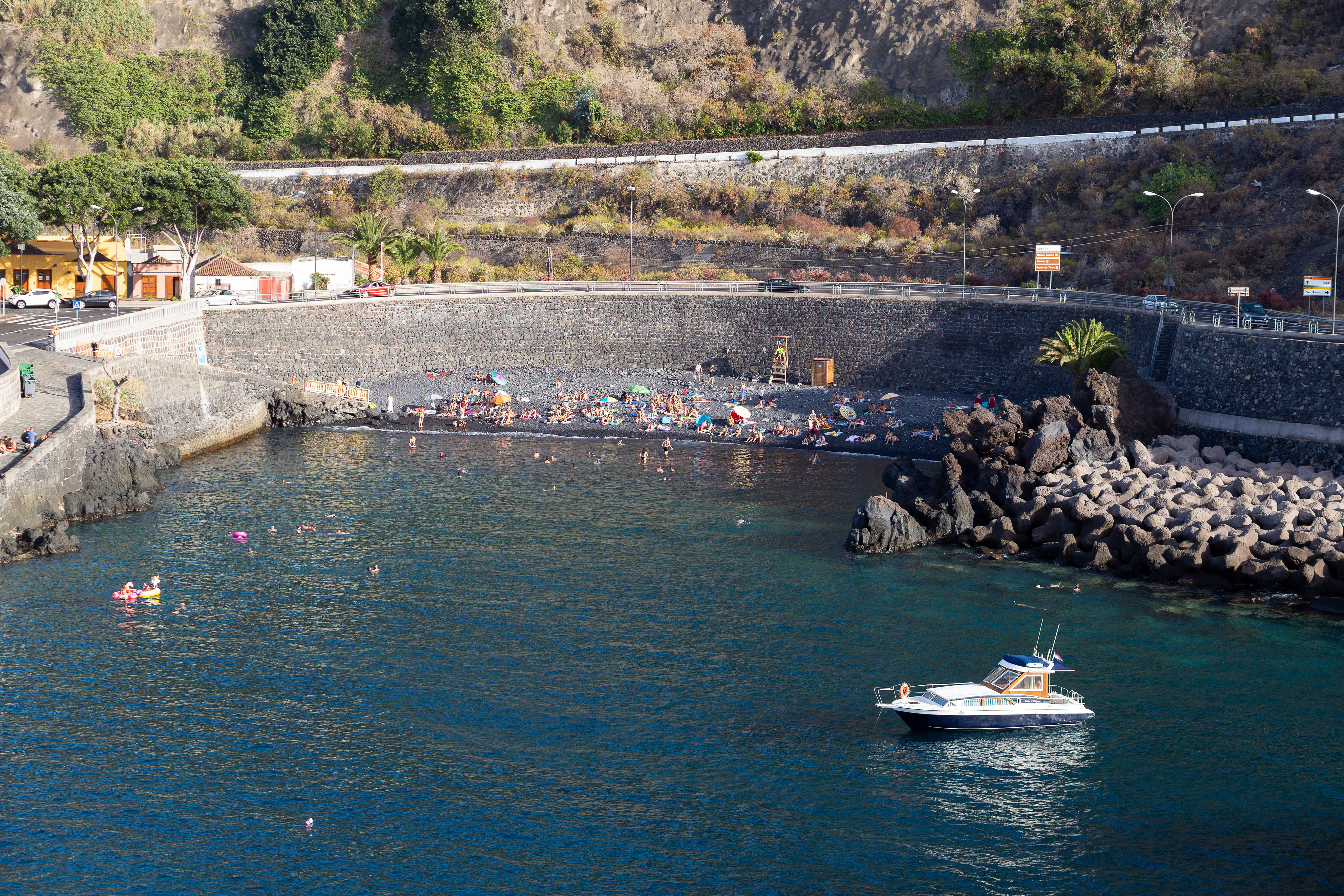 Scenic view of a black sand beach with people, blue water, a boat, and a rocky coastline.