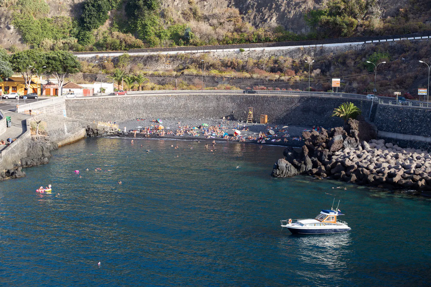 Scenic view of a black sand beach with people, blue water, a boat, and a rocky coastline.