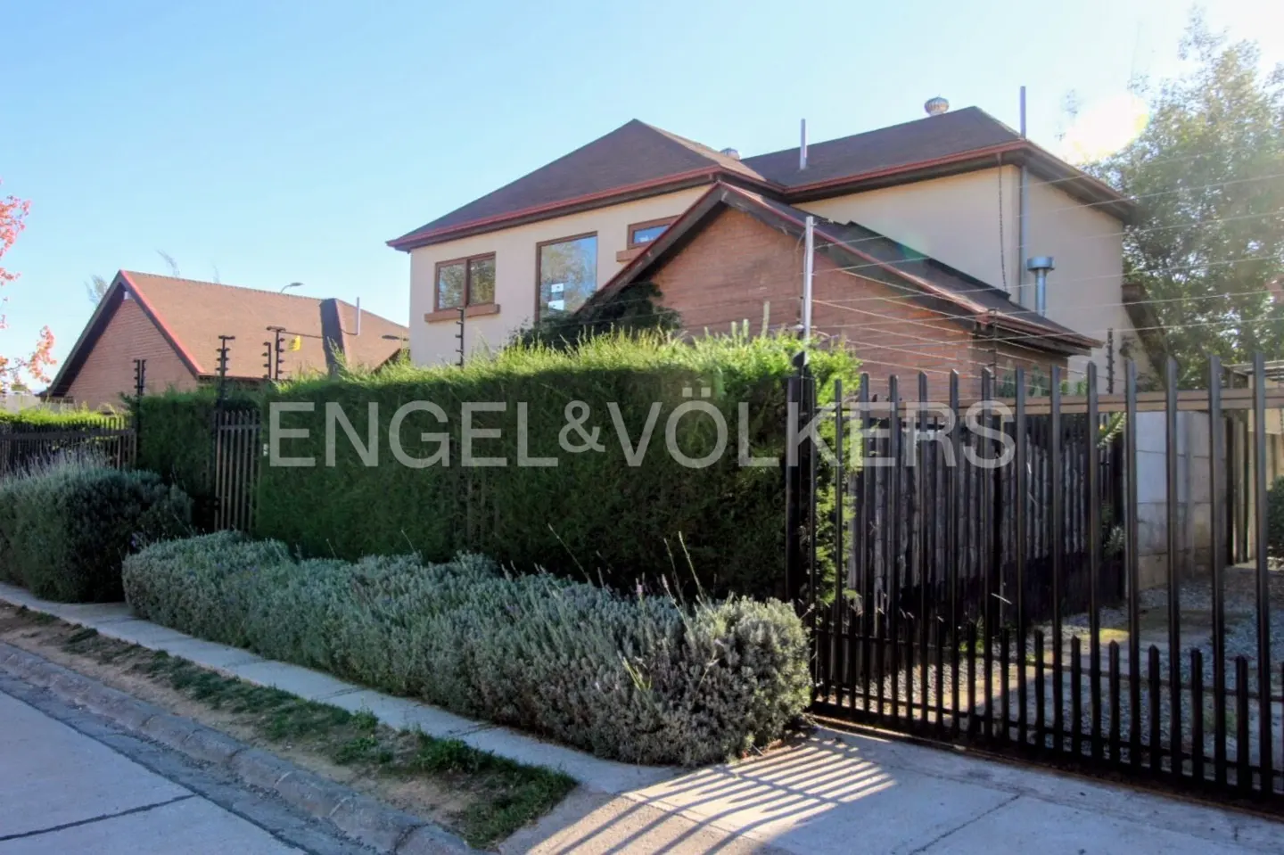 Exterior view of a two-story house with a brown roof, beige walls, and a black metal fence. The Engel & Völkers logo is visible on the hedge.