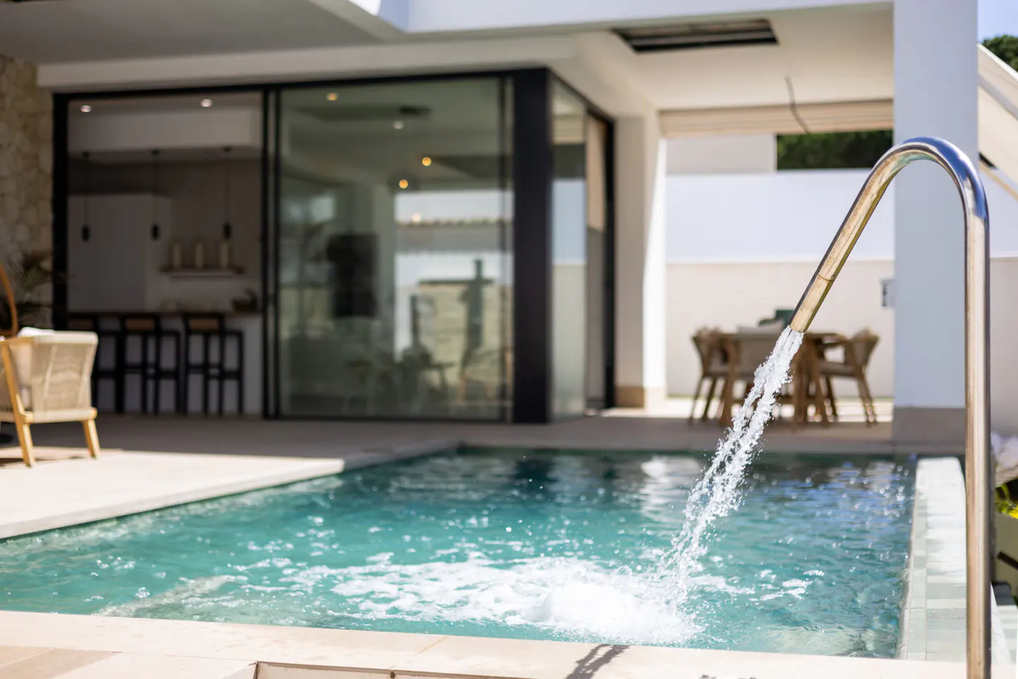A modern pool with a water feature, reflecting the outdoor dining area and kitchen through large glass doors.
