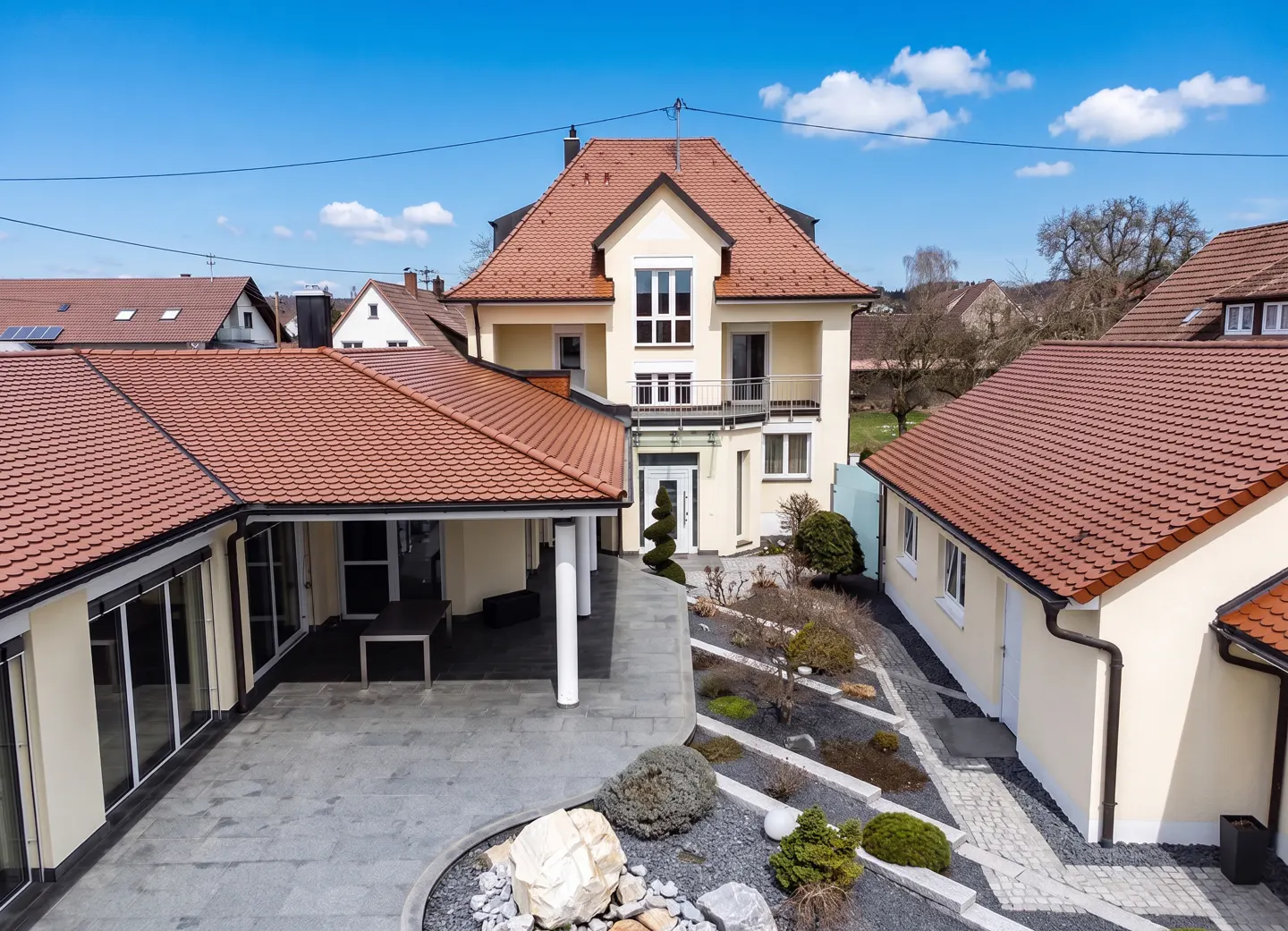 Two-story beige house with a red tile roof, a patio, and a landscaped yard under a blue sky.