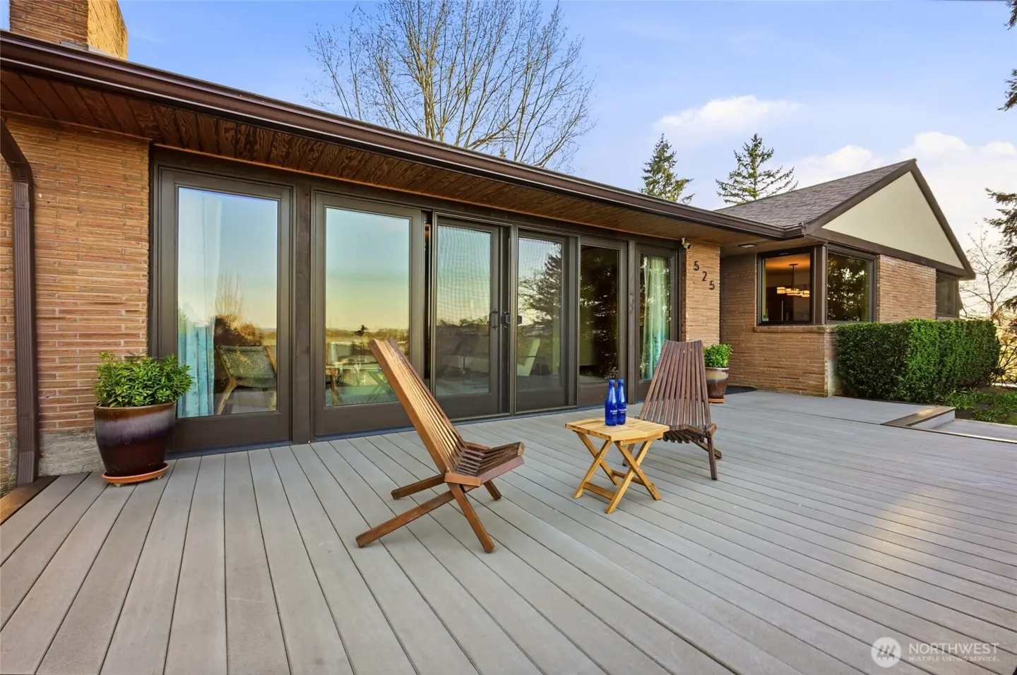Exterior view of a brick house with a gray deck, two wooden chairs, and a small table with blue bottles. Large sliding glass doors lead inside.