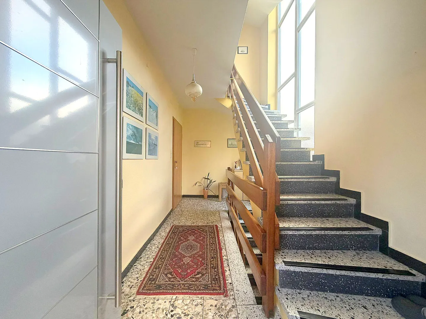 Hallway with stairs, a red rug, and a white door. The walls are yellow and decorated with framed pictures. Natural light streams in from a window.