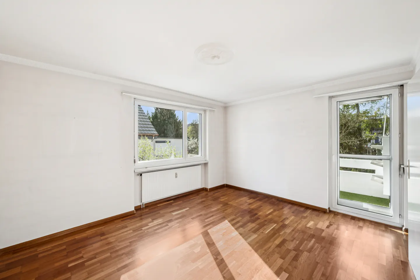 Bright, empty room with hardwood floors, white walls, a window with a radiator, and a glass door to a balcony.