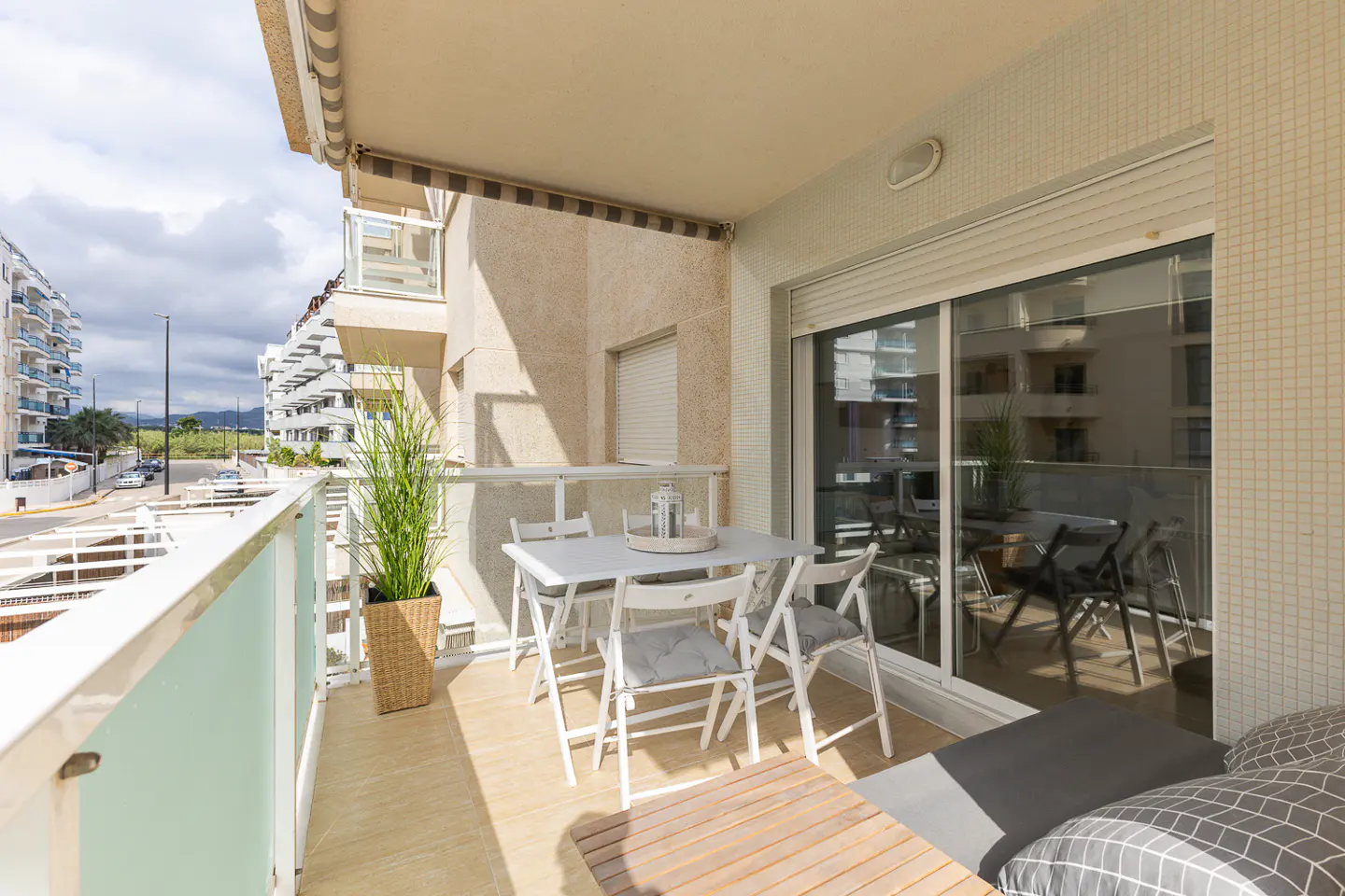 A bright apartment balcony with a dining set, a tall potted plant, and a view of a street and neighboring buildings.