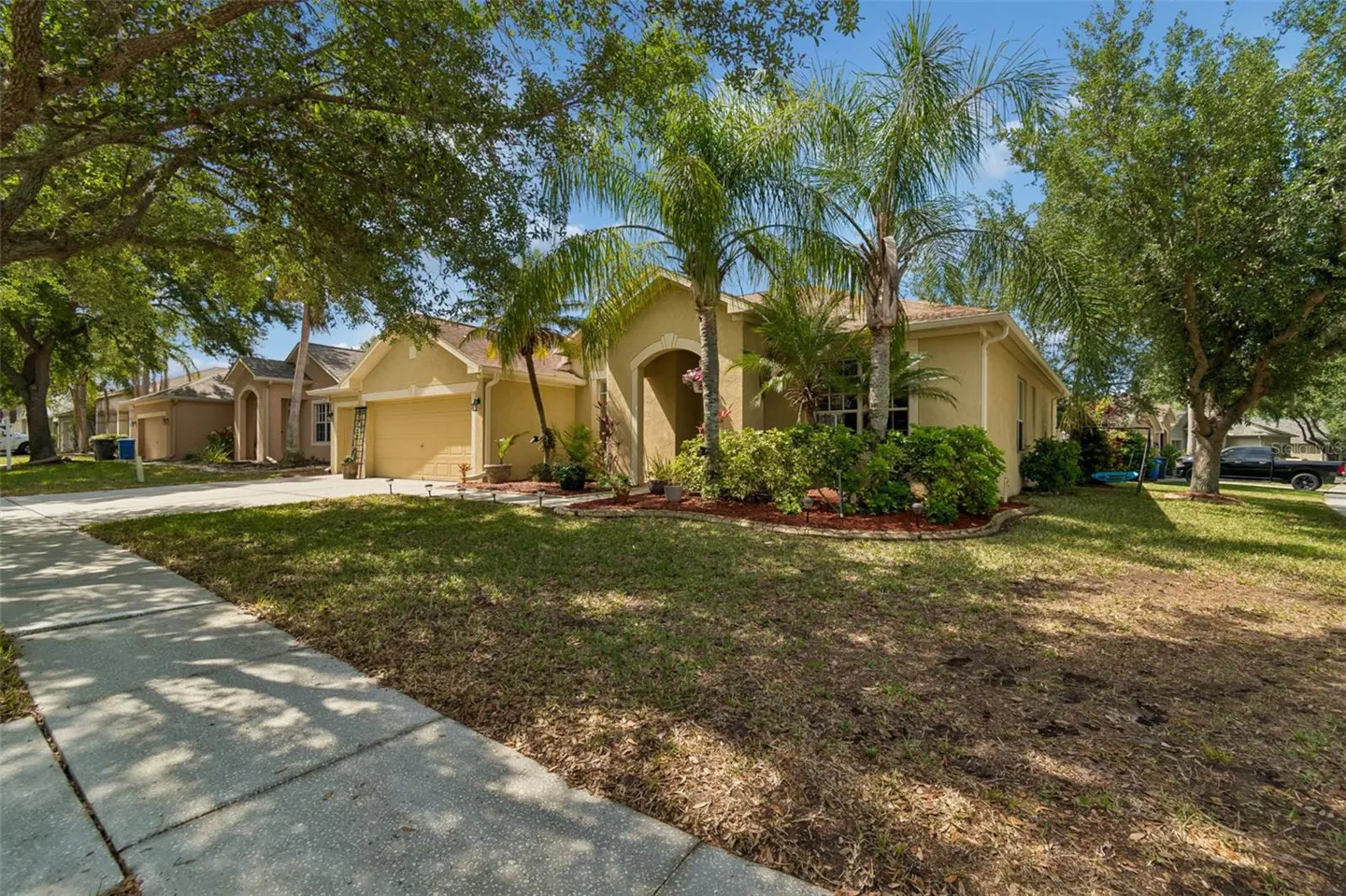 Tan single-story house with a two-car garage, palm trees, and green lawn on a sunny day.