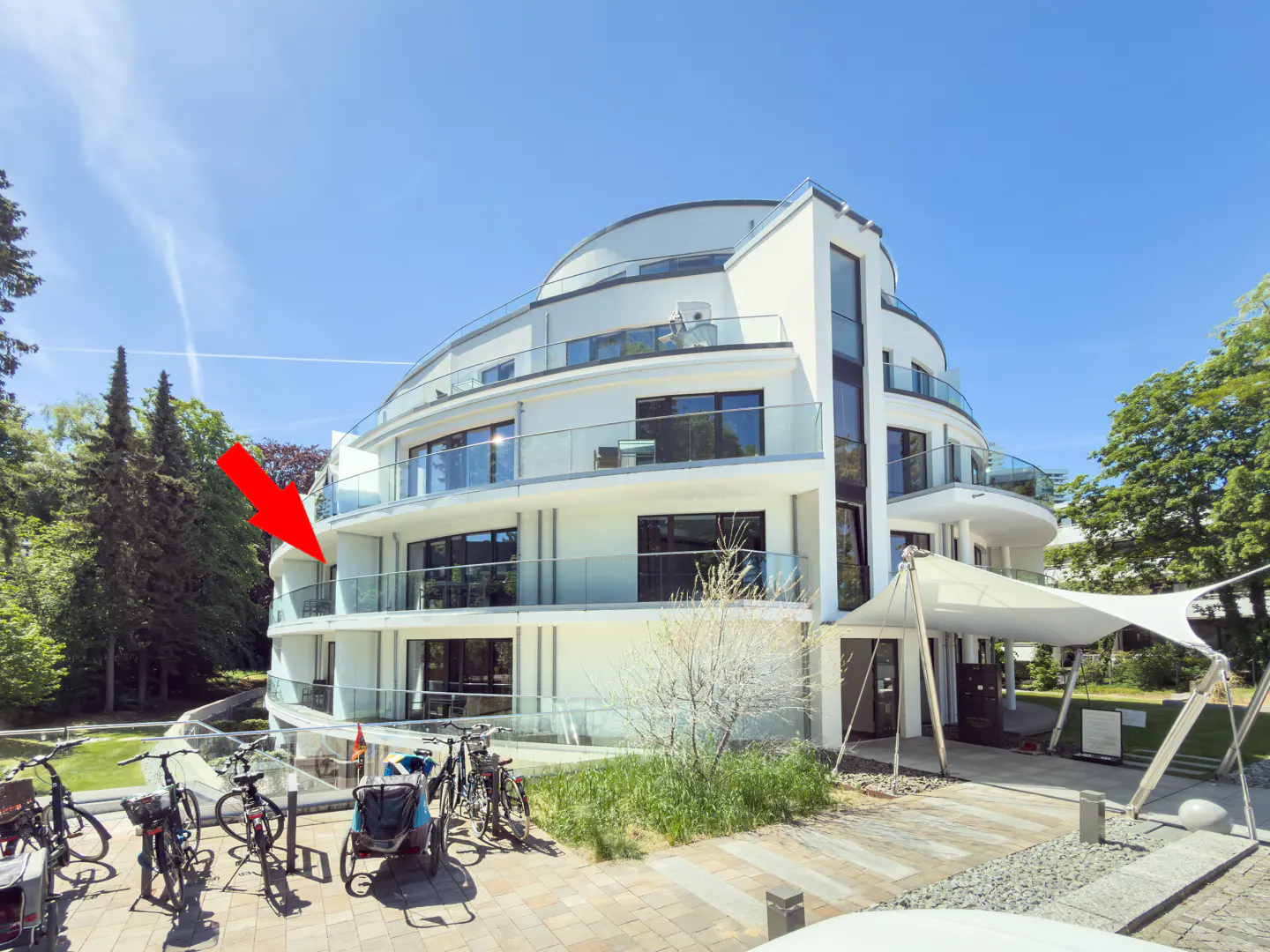 Modern white multi-story apartment building with curved balconies, a red arrow pointing to a unit, and bikes in the front.