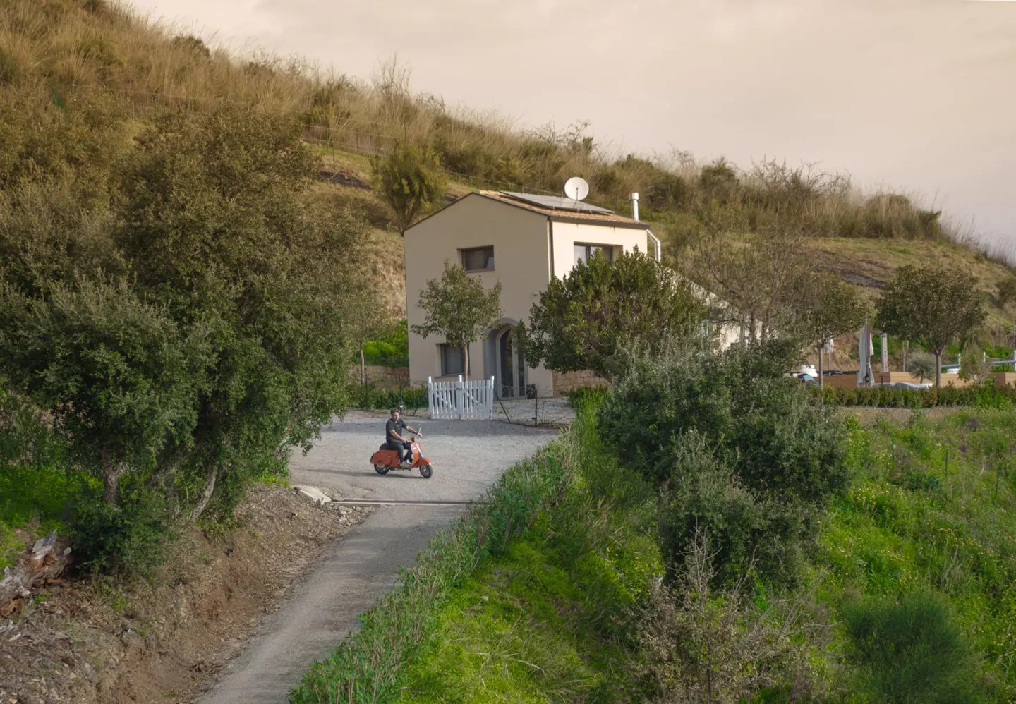 A man rides an orange scooter on a driveway leading to a modern, light-colored house on a grassy hillside.