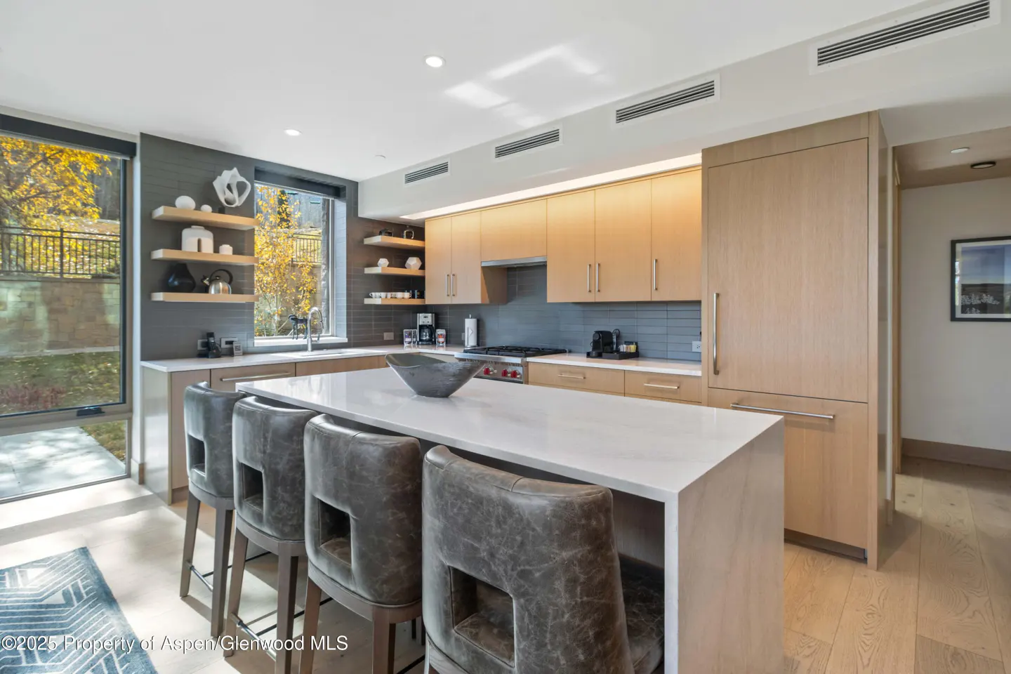 Modern kitchen with light wood cabinets, white quartz island, four grey leather bar stools, and large windows with views.