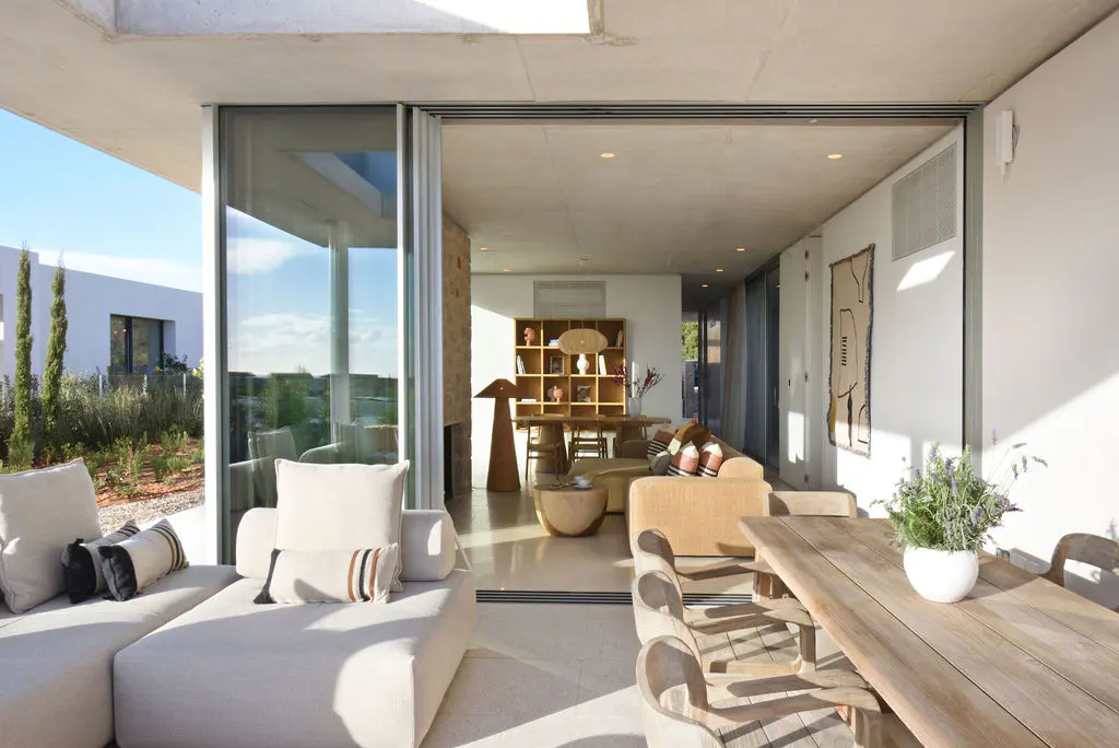 Open-concept living space with a white sectional sofa, wooden table and chairs, and a bookshelf in the background. Large sliding glass doors lead to an outdoor area.