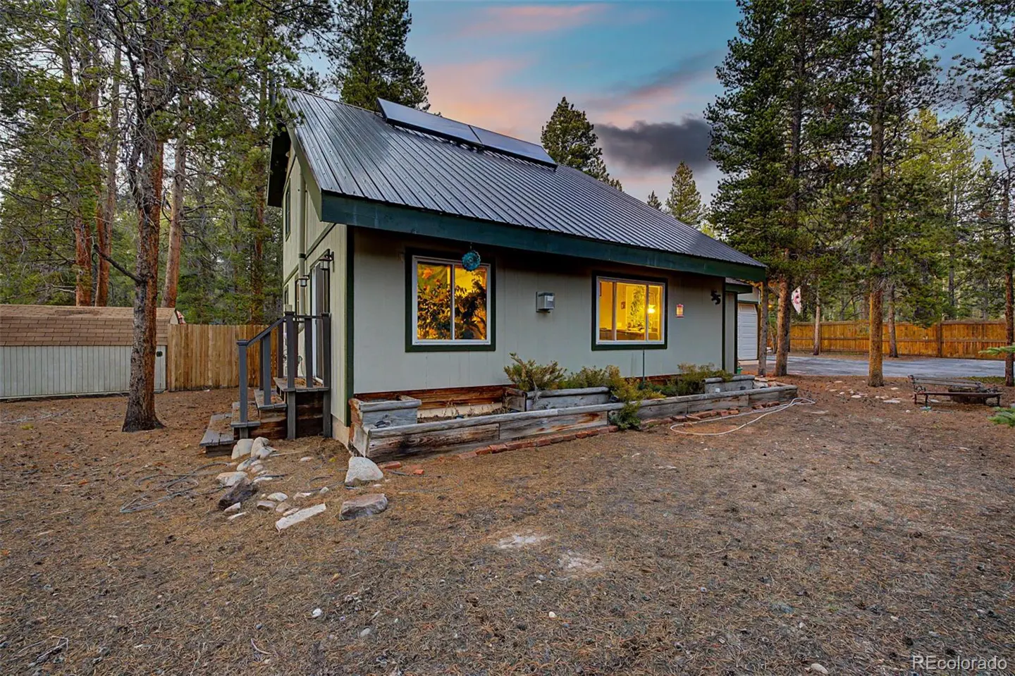 Exterior view of a light green house with a metal roof and solar panels, surrounded by tall pine trees. Two windows glow with warm light.
