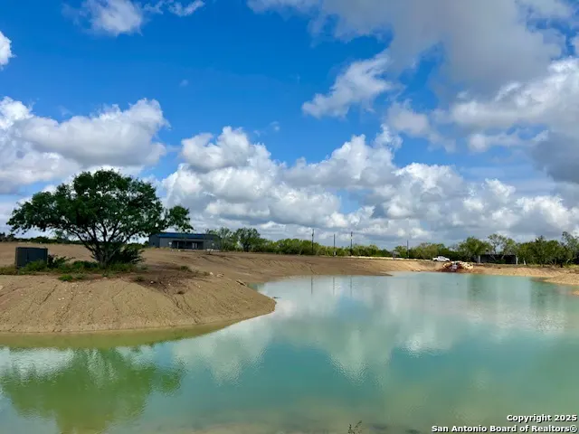 A scenic view of a pond reflecting a blue sky with fluffy white clouds. A tree and a building are visible on the sandy bank.