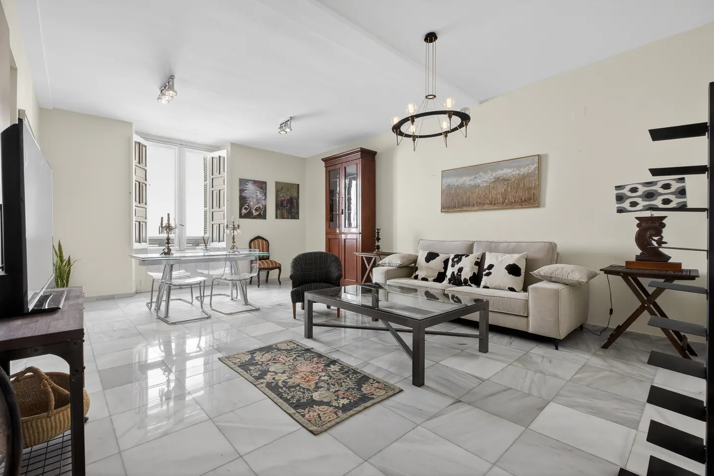 Living room with marble floors, a beige sofa with cow-print pillows, a glass coffee table, and a dining area with clear chairs.