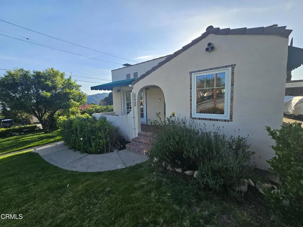 A tan stucco house with a brown tile roof, arched entry, and a blue awning. Green lawn and landscaping in front.
