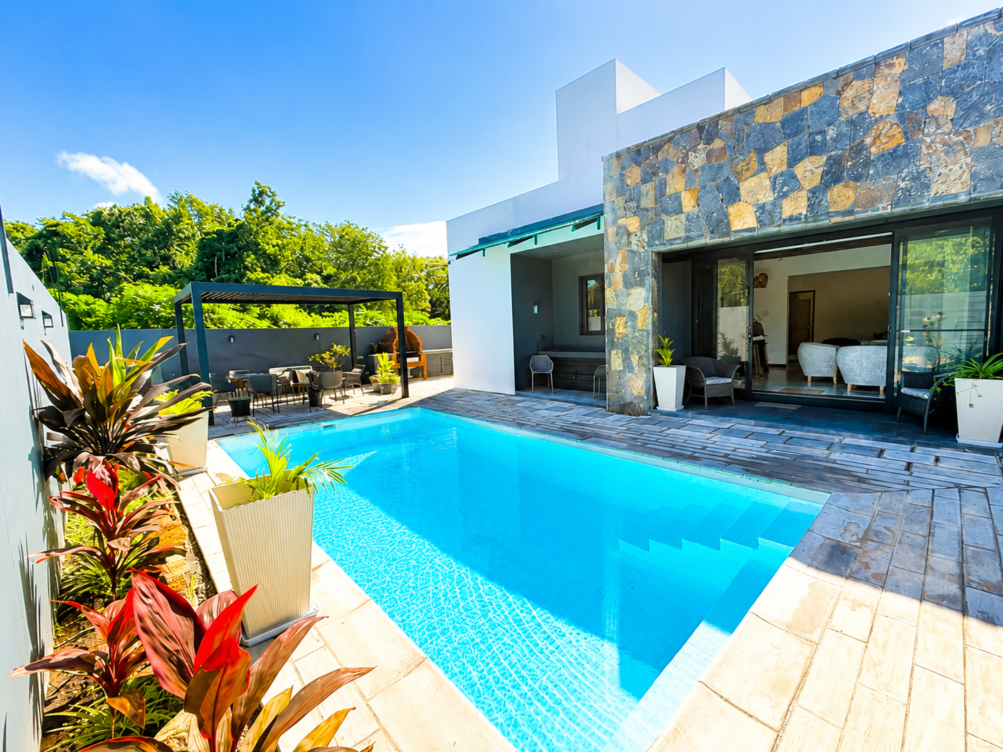 Modern home with a bright blue swimming pool, stone accent wall, patio furniture, and lush tropical landscaping.