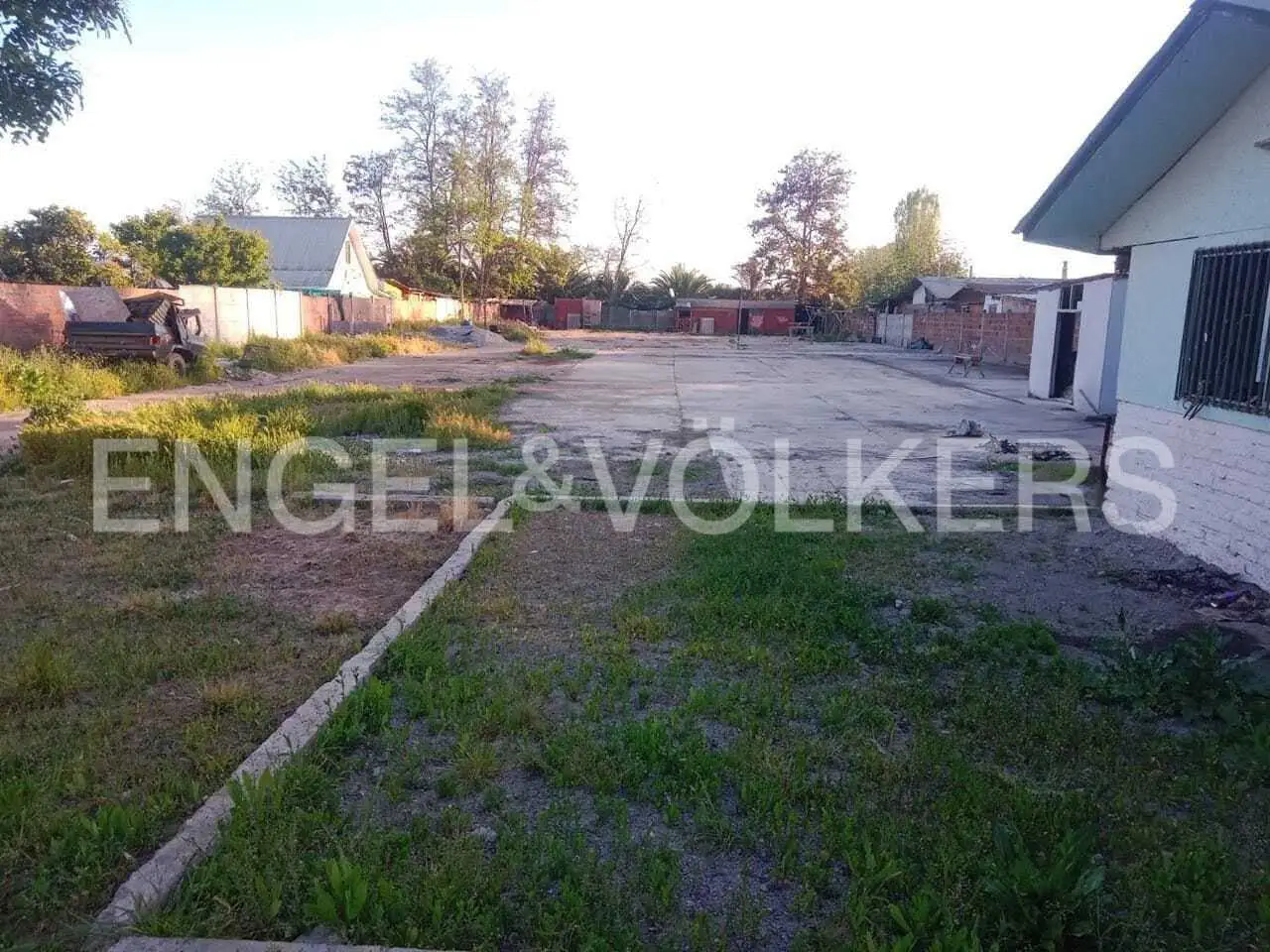 Wide shot of a large, empty concrete lot with grass patches and a building on the right. Trees and other buildings are in the background.