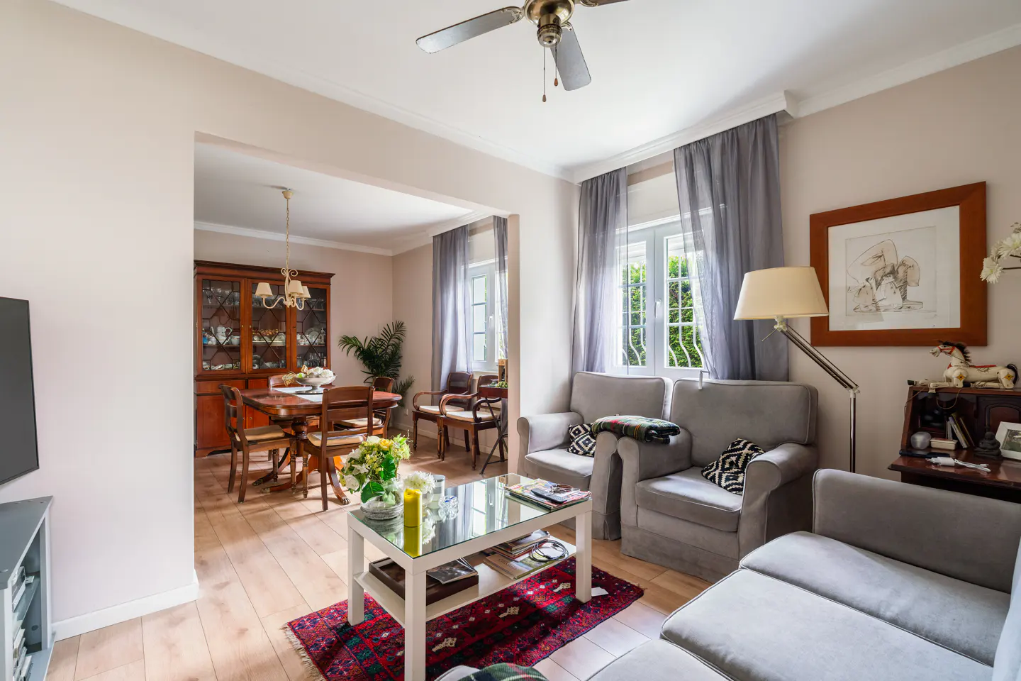 A bright, open-concept living and dining room with hardwood floors, gray sofas, and a glass coffee table. A dining table and chairs are visible in the background.