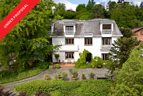A white, multi-story house with a gray roof sits on a hill surrounded by green trees. A red banner reads "UNDER PROPOSAL."