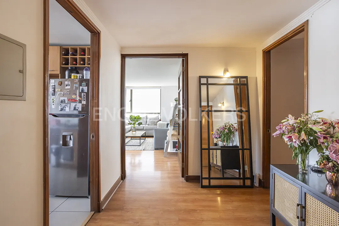 Hallway with wood floors, white walls, and wood trim. A stainless steel refrigerator is visible through a doorway. A mirror and flowers sit on a black cabinet.