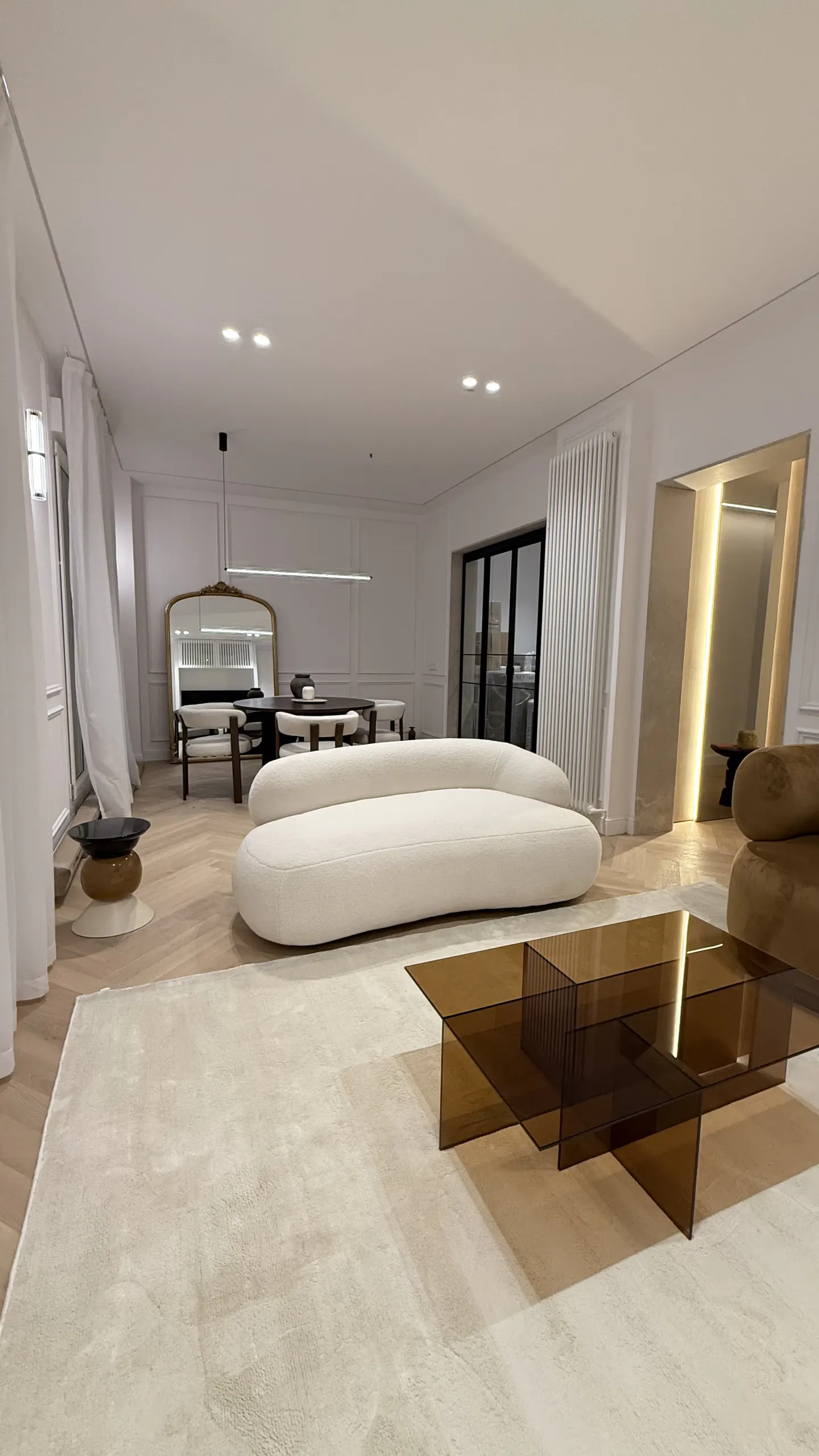 Modern living room with a curved white sofa, amber glass coffee table, and light wood floors in a bright, open space.