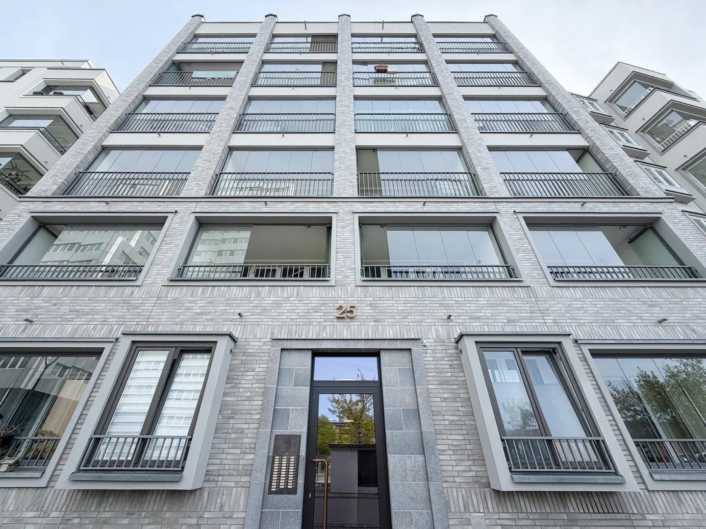 Exterior view of a tall, gray brick apartment building with balconies and the number 25 above the entrance.