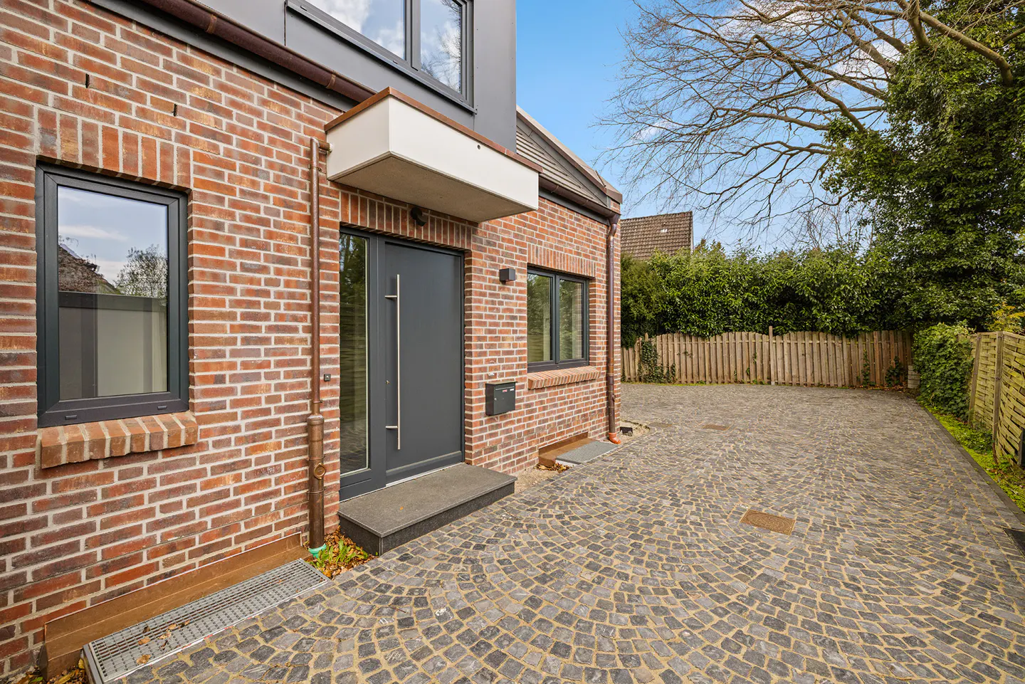 Brick house exterior with gray door, windows, and cobblestone driveway. Green bushes and a wooden fence are in the background.