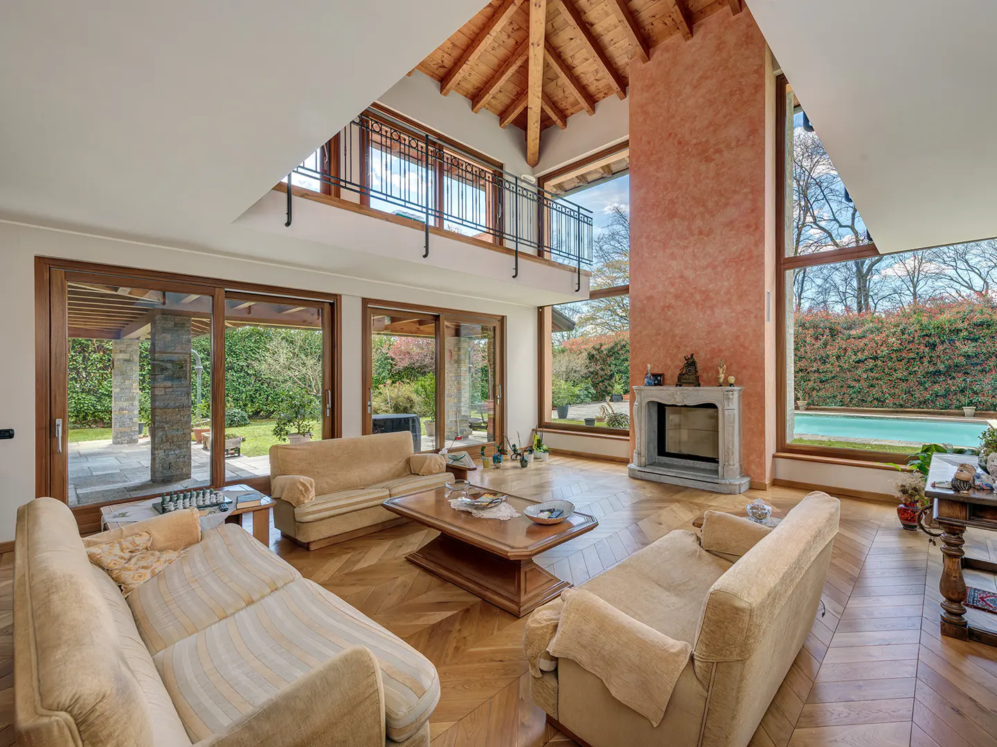 Spacious living room with high vaulted wood ceiling, stone fireplace, beige sofas, and large windows overlooking a pool.