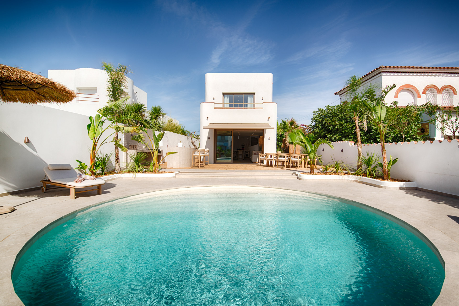 A round swimming pool in a sunny courtyard with a white modern house, lounge chair, and tropical plants under a blue sky. [AI-generated text]